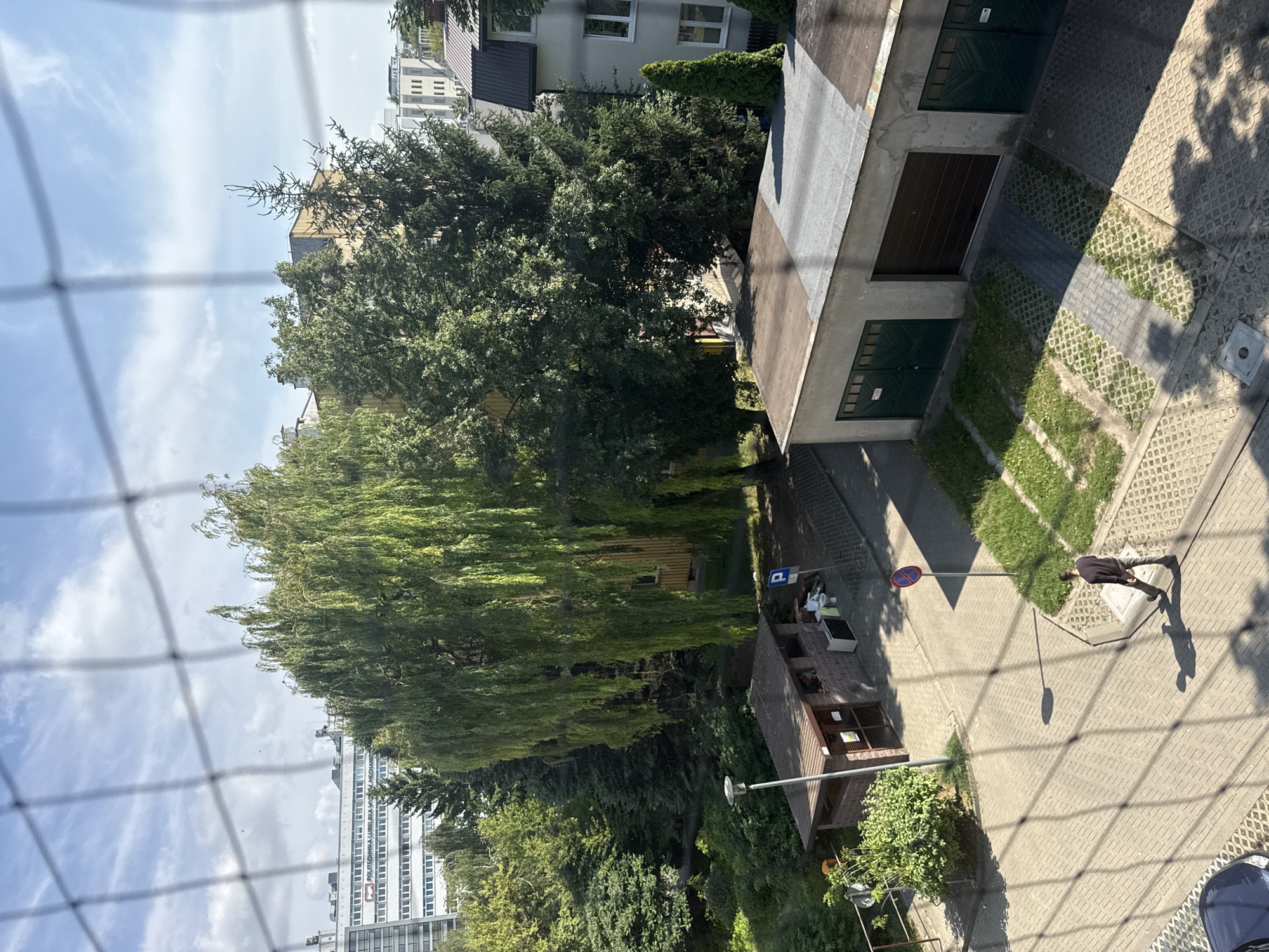 This is a high-angle, medium shot taken from a building looking out over a courtyard area in the city of Lublin, Poland. A prominent weeping willow tree dominates the left side of the frame, its long, green branches hanging down. Behind the willow and to the right are various other trees, including a tall evergreen.
In the foreground, a wire mesh fence is visible, casting a grid-like shadow across the scene. A man in a dark long-sleeved shirt and light-colored pants is walking across a paved area in the lower center of the image, his shadow cast by the sun.
To the left, there is a small brick structure with a brown roof, appearing to be some kind of utility shed or kiosk, with two doors and an open window. A street lamp stands next to it. Further back, a tall, white, multi-story building with many windows is visible, with the text "POLITECHNIKA LUBELSKA" and "WYDZIAL MECHANICZNY" visible on its facade.
In the center of the courtyard, there are several grassy patches and a paved walkway. To the right, a row of garages with dark brown and green doors is visible. A blue sign with a white 'P' and a red circle with a diagonal line indicates a parking area. The sky is blue with scattered white clouds. The overall impression is an urban scene with a mix of residential and institutional buildings, and a focus on green spaces and trees.