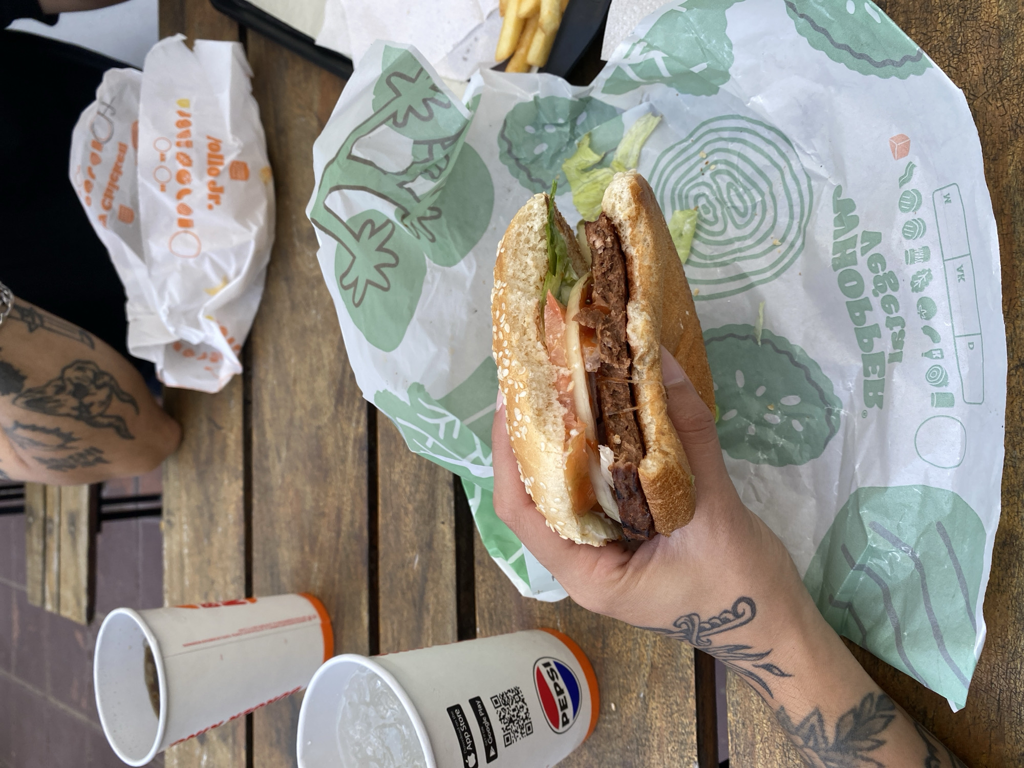 A person with tattoos is holding a burger on a wrapper with green tomato shapes in their right hand. A cup of Pepsi is next to the burger, as is another empty cup. There is a wrapper for Jollo Jr. chicken, and some fries, on the table, which is made of horizontal wooden slats. The location is identified as Monte Grande, Argentina.