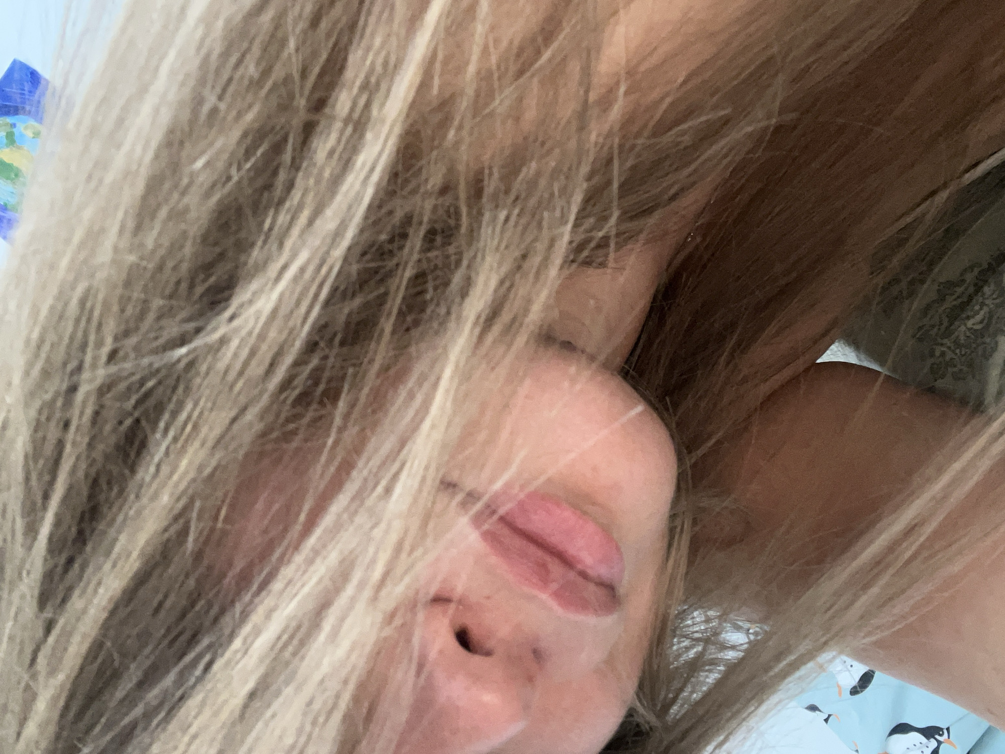 Close-up of a person's face with light blonde hair partially obscuring it. Visible features include the nose, lips, and partially visible eye. The person is lying down on a blue sheet with penguin motifs. The location and city of Budva, Montenegro cannot be determined based on the image.