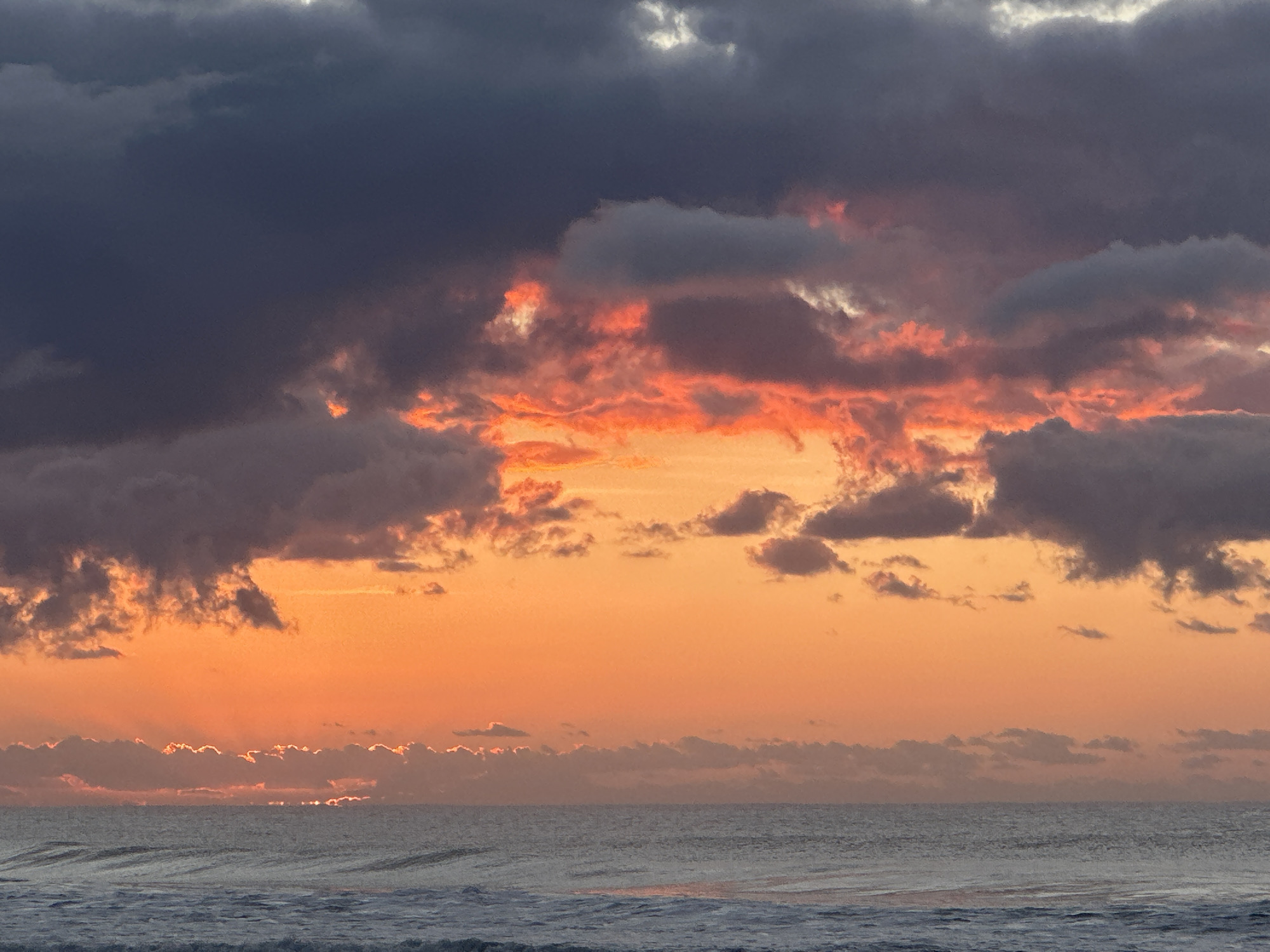 The image depicts a seascape at sunrise over Mylestom, Australia. The sky is filled with dramatic clouds, with a large portion of dark grey clouds dominating the upper left. In the center and right, the clouds are illuminated by the rising sun, creating vibrant shades of orange and pink, with streaks of light breaking through. A band of smaller, illuminated clouds stretches across the horizon. Below the sky, the ocean is visible, with gentle waves reflecting the colors of the sky. The overall scene conveys a sense of natural beauty and the start of a new day. There are no people, subjects, objects, or actions depicted in the image.