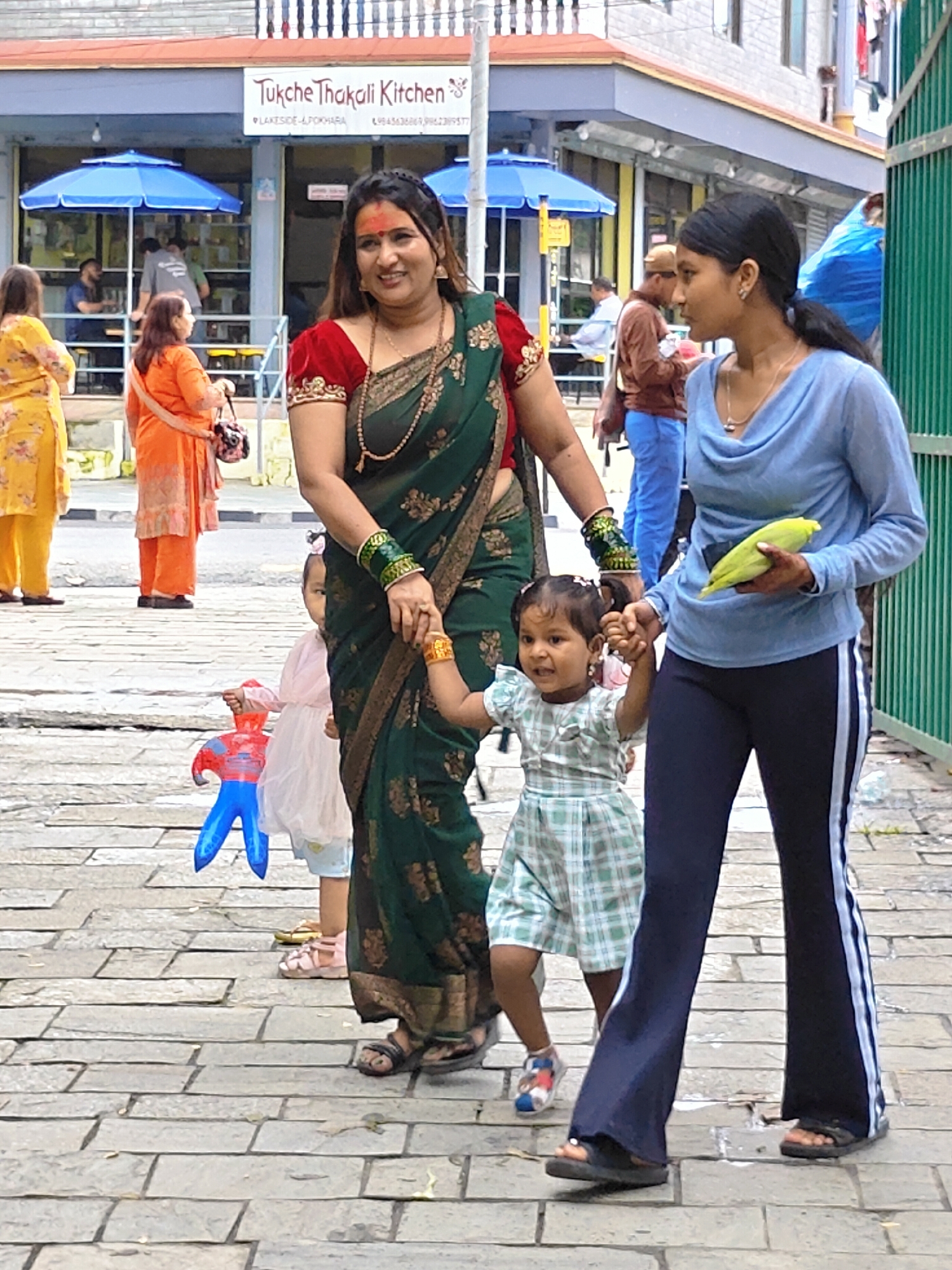 In Pokhara, Nepal, two women and two children are walking on a paved sidewalk. The woman on the left, dressed in a green sari with gold embroidery, has a red tika on her forehead and is wearing a beaded necklace and bracelets. She is holding the hand of a young girl in a green and white checkered dress. The child is holding a blue and red inflatable Spiderman toy. Beside the child, another young girl in a pink dress is visible. The woman on the right, wearing a light blue long-sleeved shirt and dark blue pants with white stripes, is holding a yellow cob of corn in her right hand. The background shows a building with a sign that reads "Tukche Thakali Kitchen." Blue umbrellas are visible outside the establishment. The ground is paved with rectangular stones.