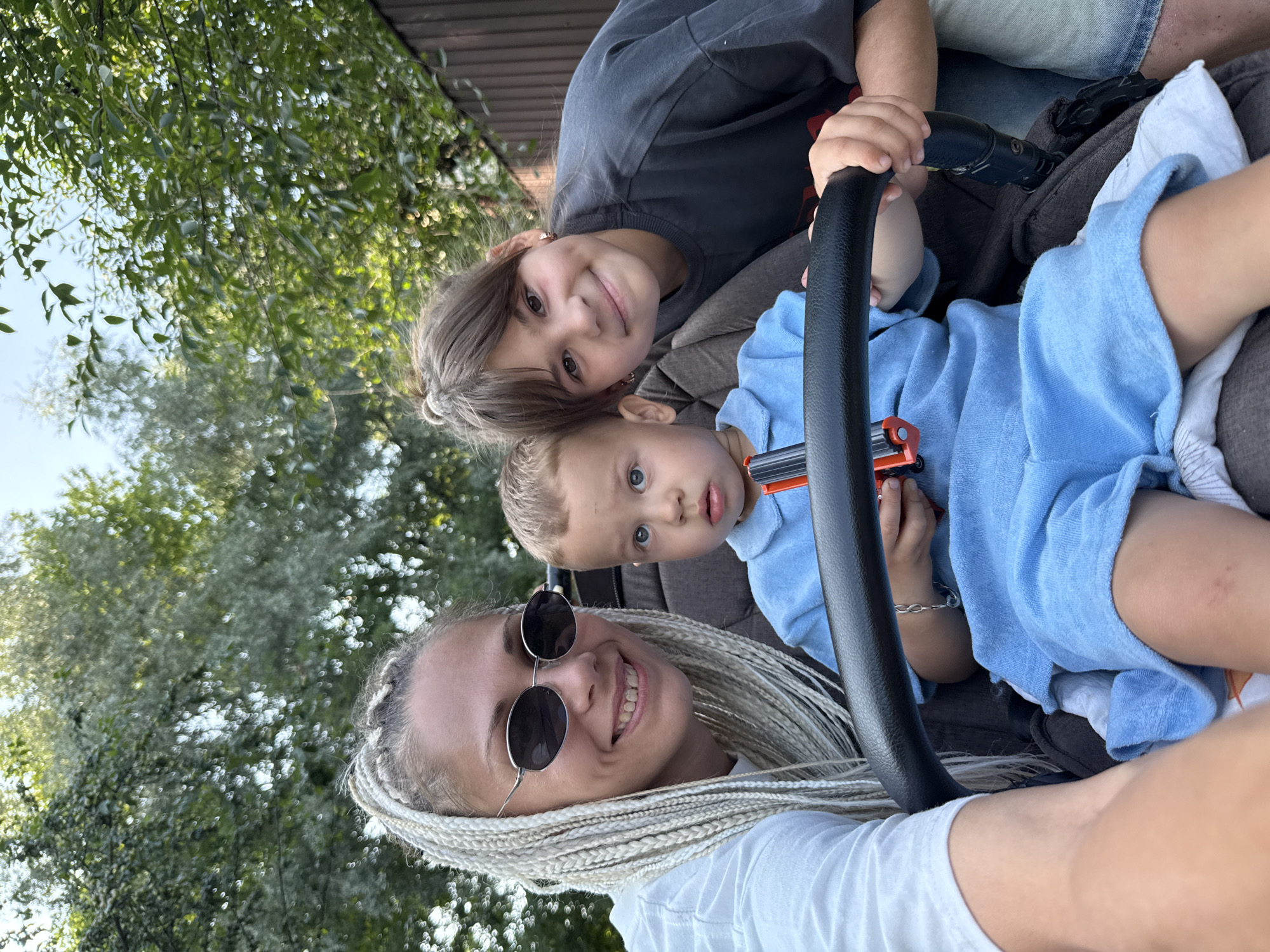 In Rusanovo, Russia, a woman with blonde braided hair and sunglasses smiles at the camera, seated in the front of a stroller. Beside her, a young girl with brown hair also smiles at the camera. In the stroller's seat, a young boy wearing a blue outfit looks towards the camera, holding a toy. The background is a natural setting with trees and a wooden fence, suggesting an outdoor location.