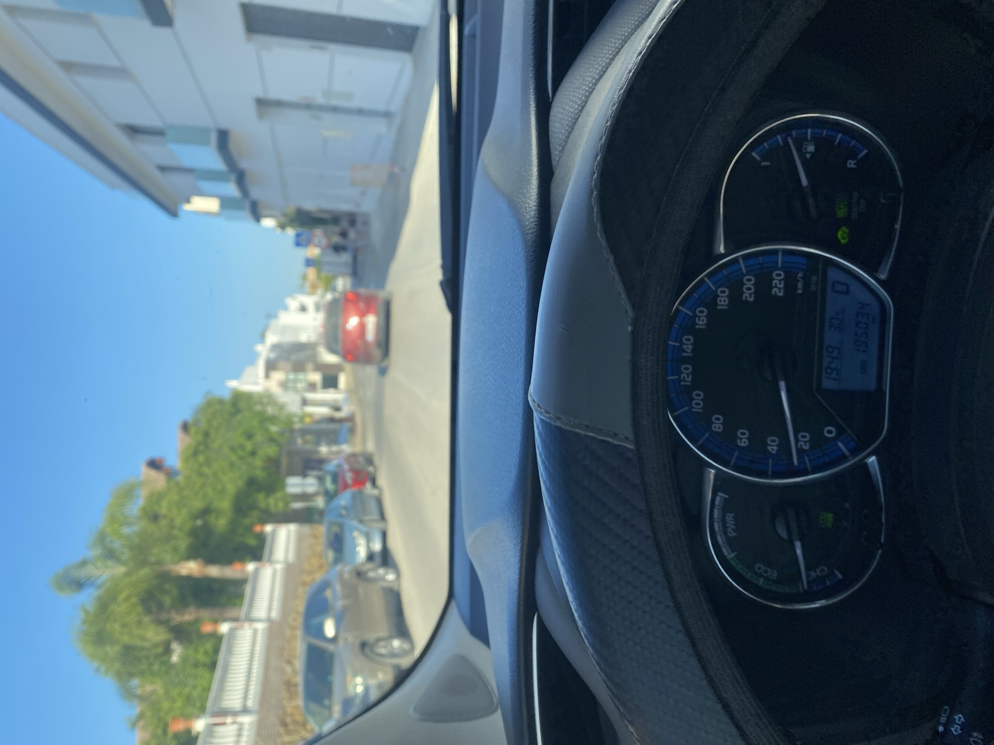 The image is taken from the driver's seat of a car, looking forward through the windshield. The dashboard is visible in the foreground, with a steering wheel covered in black and grey textured material. To the right of the steering wheel is the car's instrument cluster, which includes a speedometer, an ECO/PWR gauge, a fuel gauge, and a digital display showing the time as 19:49 and the odometer reading as 195034. The car is driving on a street lined with white buildings with balconies, consistent with the architecture found in Estepona, Spain. Trees, including palm trees, are visible on the left side of the street, along with a white fence and terracotta-topped pillars. Several cars are parked along the street, and a red car is in front of the viewpoint car. The sky is clear and blue, indicating daytime. The scene depicts a typical urban street in a sunny climate.