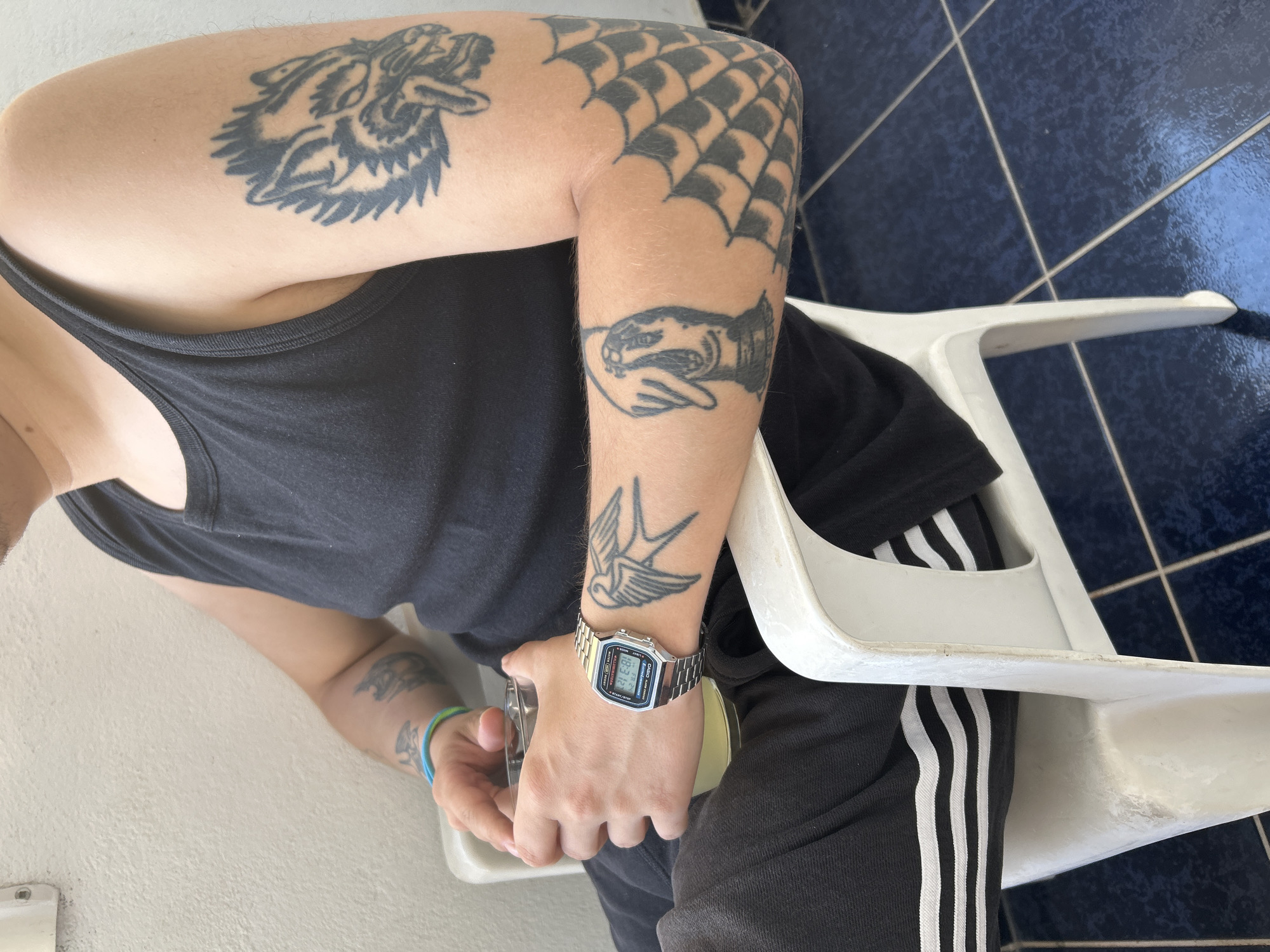 A person wearing a black tank top and black sweatpants with white stripes sits in a white plastic chair on a blue tiled floor. The person is holding a clear glass with a light yellow beverage. A silver digital watch is worn on the left wrist. The person has several tattoos on their arms, including a wolf head on the upper left arm, a bird on the left forearm, and a web-like pattern on the right forearm. The setting appears to be outdoors, possibly on a balcony or patio.