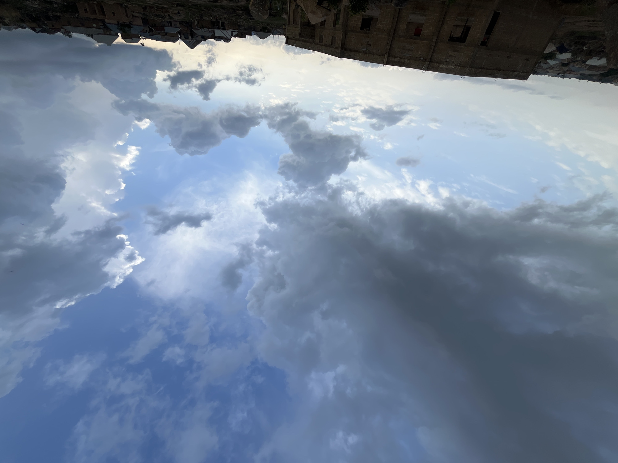 The image depicts a cloudy sky above Jos, Nigeria. The sky is filled with a mix of white and dark grey cumulus clouds, suggesting varied weather conditions. In the top portion of the image, upside down, there is a glimpse of buildings, possibly houses, indicating an urban or residential area. Some buildings appear to be under construction or in a state of disrepair. There are no people visible in the frame. The environment suggests it is daytime, based on the illumination, and the setting is a blend of sky, clouds, and urban structures. The overall scene is a landscape view focusing on the sky and the top of the buildings.