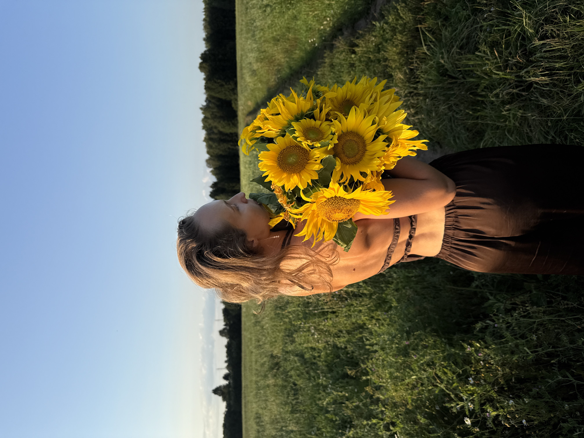 In Aksaitovo, Russia, a woman stands in a field of green grass, holding a bouquet of sunflowers. She is wearing a dark brown dress with an open back. The sun is setting, casting a warm glow on the scene. The sky is clear and blue, with a few clouds in the distance. The field is bordered by a forest.