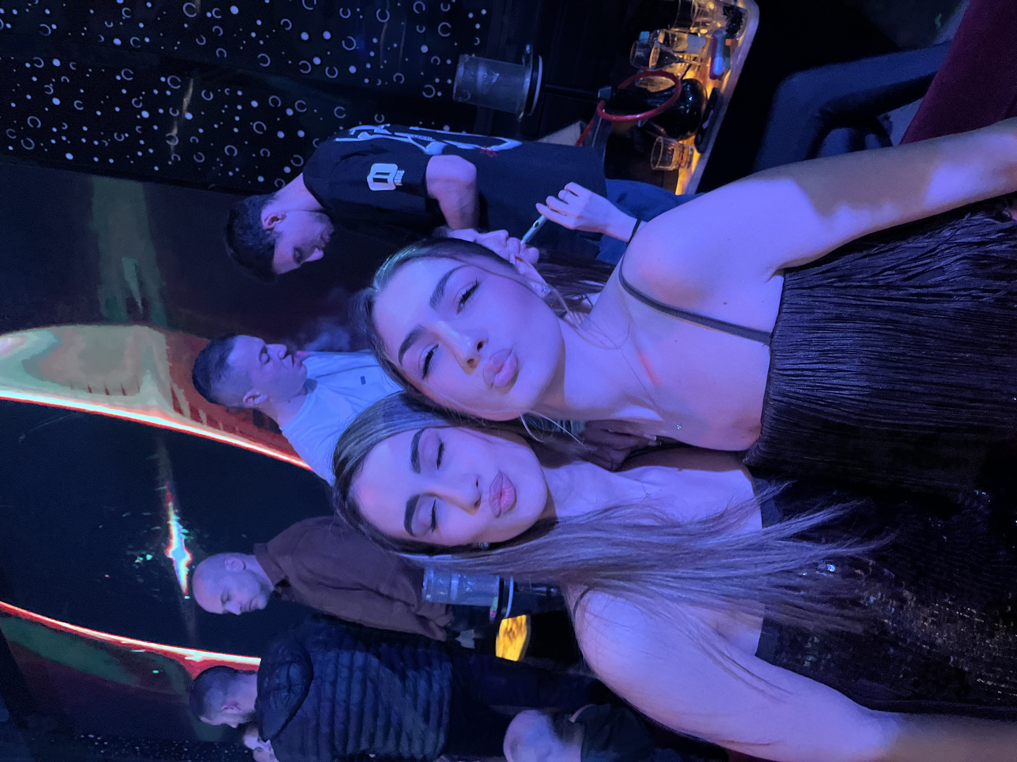 Two young women are posing closely together for a selfie. Both have their lips pursed as if giving a kiss. Behind them, several people are standing or sitting, including three men with shaved heads and others who are partially obscured by the dark environment. In the background, there's a table with glasses and bottles, along with what appears to be a hookah. The setting suggests a dimly lit nightclub or lounge, possibly in Burgas, Bulgaria. A shiny black textured material reflects light creating linear shapes.