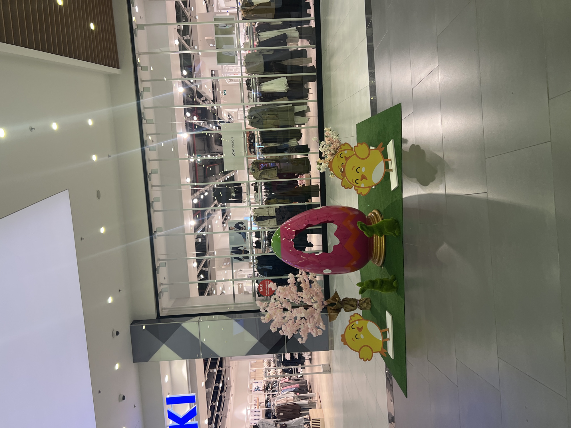 The image depicts an Easter display inside what appears to be a shopping mall in Plovdiv, Bulgaria. The display features a large, pink Easter egg with a jagged hole in the center, positioned on a green artificial grass patch. Additional elements include cartoonish chick cutouts, artificial bunny figurines, and a decorative cherry blossom branch. The display is located in front of a retail store with a glass facade showcasing clothing racks and mannequins. The storefront next to it is a blue and white KT store. The surrounding environment is a typical mall interior, with tiled floors and overhead lighting. No people are visible in the immediate foreground. The overall impression is that of an Easter-themed promotional area set up to attract shoppers within the mall.