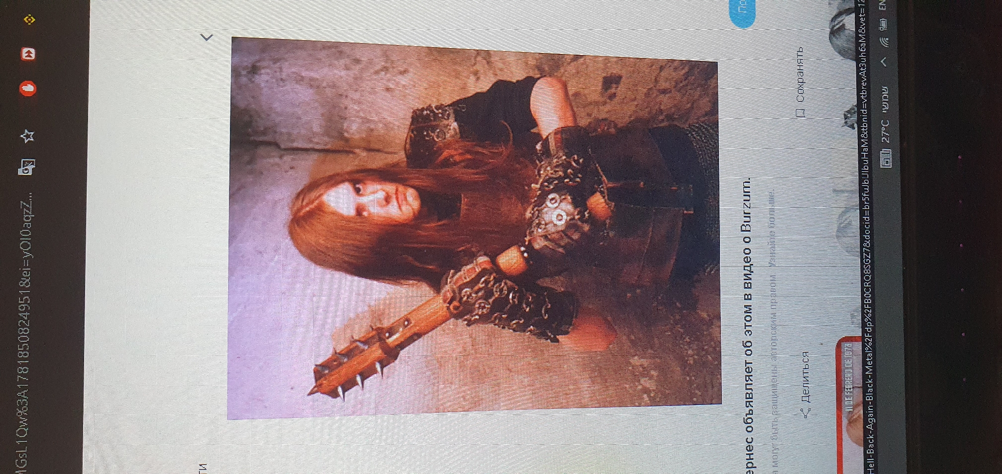 The image shows a person with long, reddish-brown hair, wearing a dark outfit and elaborate chain-like armor on their arms, holding a spiked wooden club. The person has a stern expression and is positioned against a textured, stone-like background. Below the image, a caption in Russian reads, "чернес объявляет об этом в видео о Burzum." (Chernes announces this in a video about Burzum). Further text below includes options to "Делиться" (Share) and "Сохранять" (Save). The bottom of the image displays a URL and system information indicating a temperature of 27°C and the language Hebrew ("שמשי"). The image appears to be a screenshot from a digital device, possibly a computer or phone, displaying a web page. There is no identifiable information or imagery in the provided image that specifically places the scene in the city of Bat Yam, Israel.