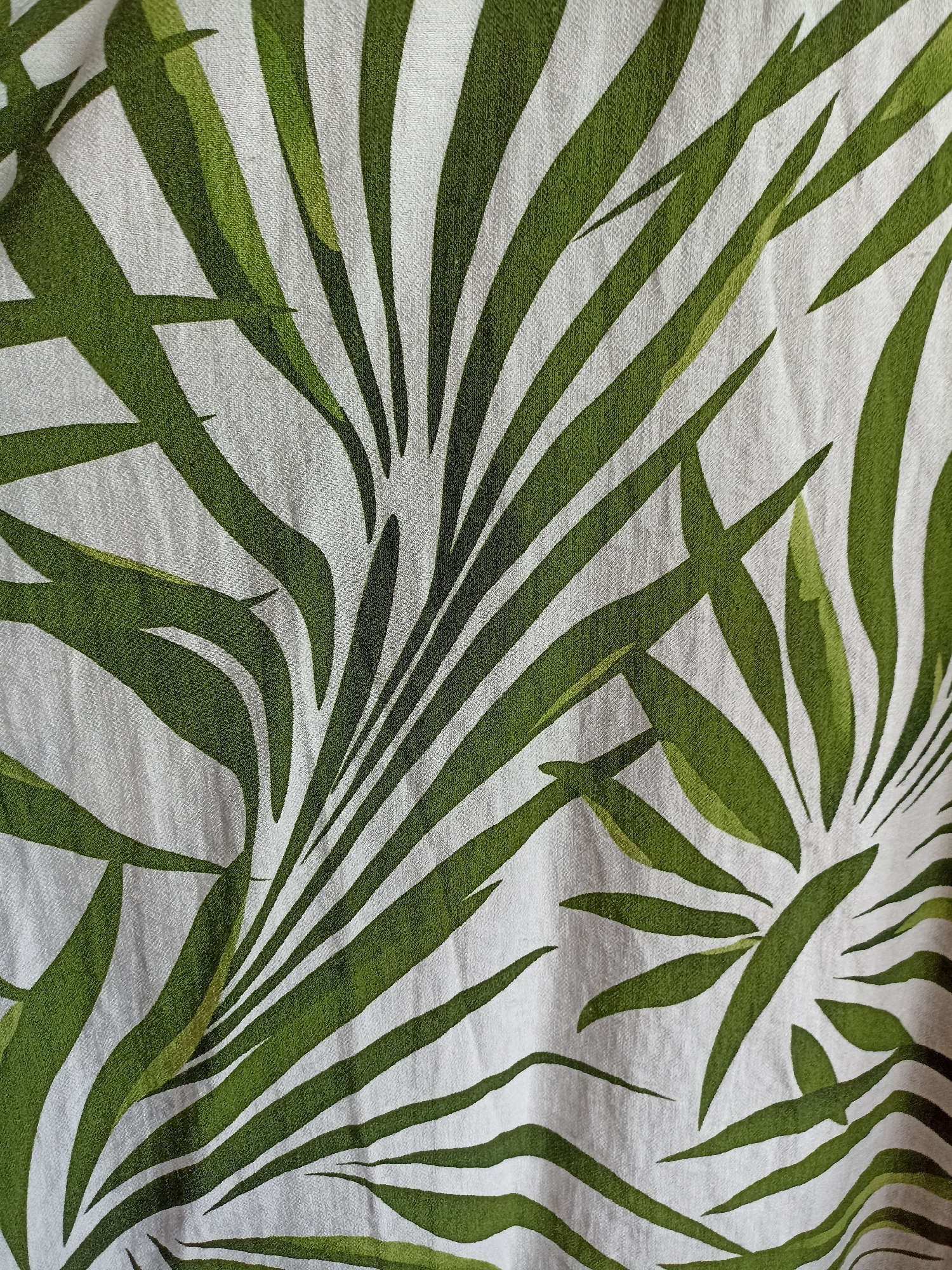 The image is a close-up, full-frame shot of a white fabric with a green palm leaf pattern. The leaves are depicted in various shades of green, with some appearing darker and others lighter, suggesting depth and shading. The fabric has a visible texture, with a slight crinkle or weave pattern. The image is an artistic representation of foliage on textile and does not depict any specific location in Novosibirsk, Russia or any identifiable subjects, actions, or interactions.