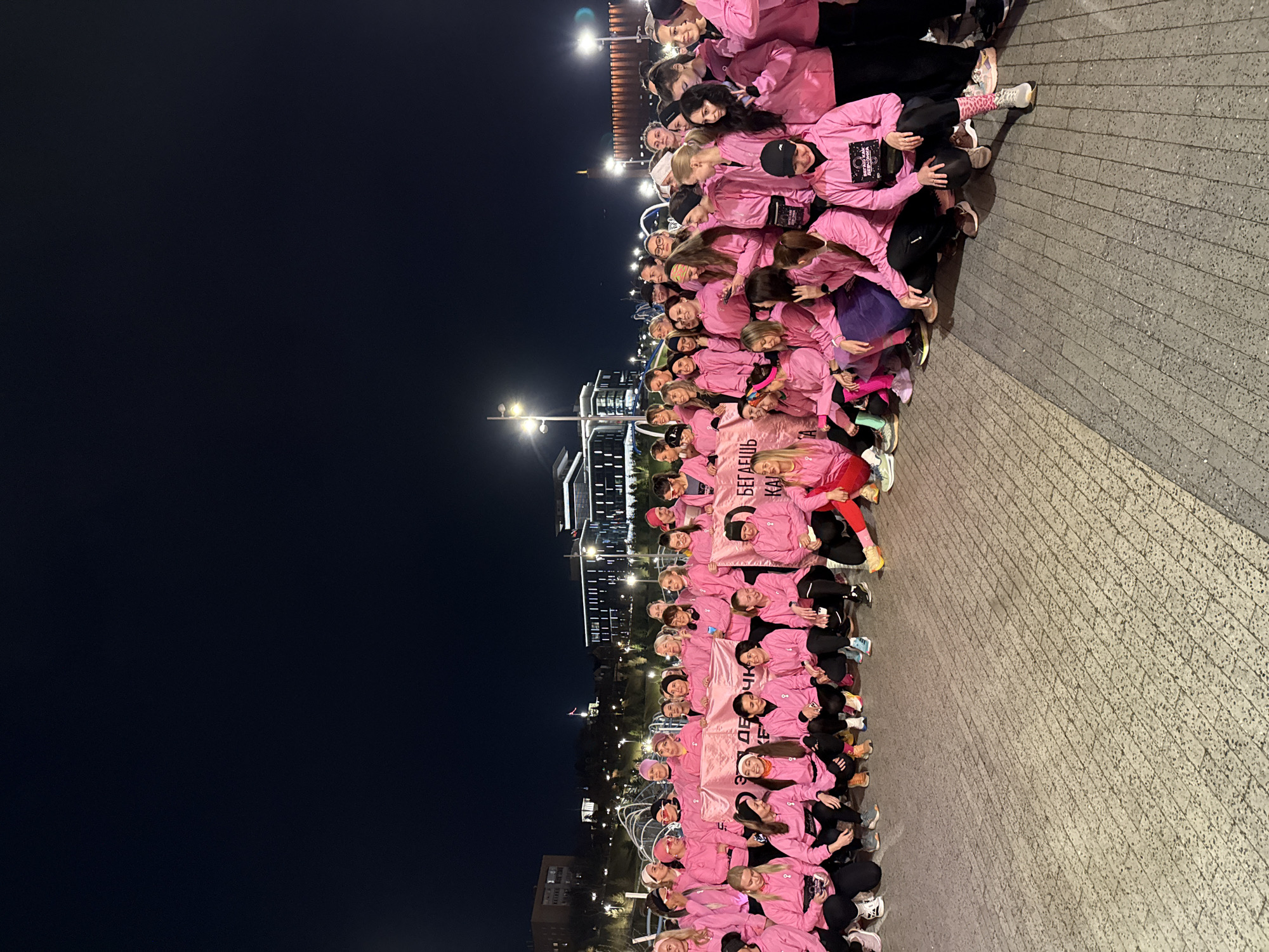 At night in Kazan, Russia, a large group of people wearing pink tops pose for a photo on a stone-paved area. The group is holding a pink banner with Cyrillic writing. In the background, buildings and streetlights are visible against a dark sky.