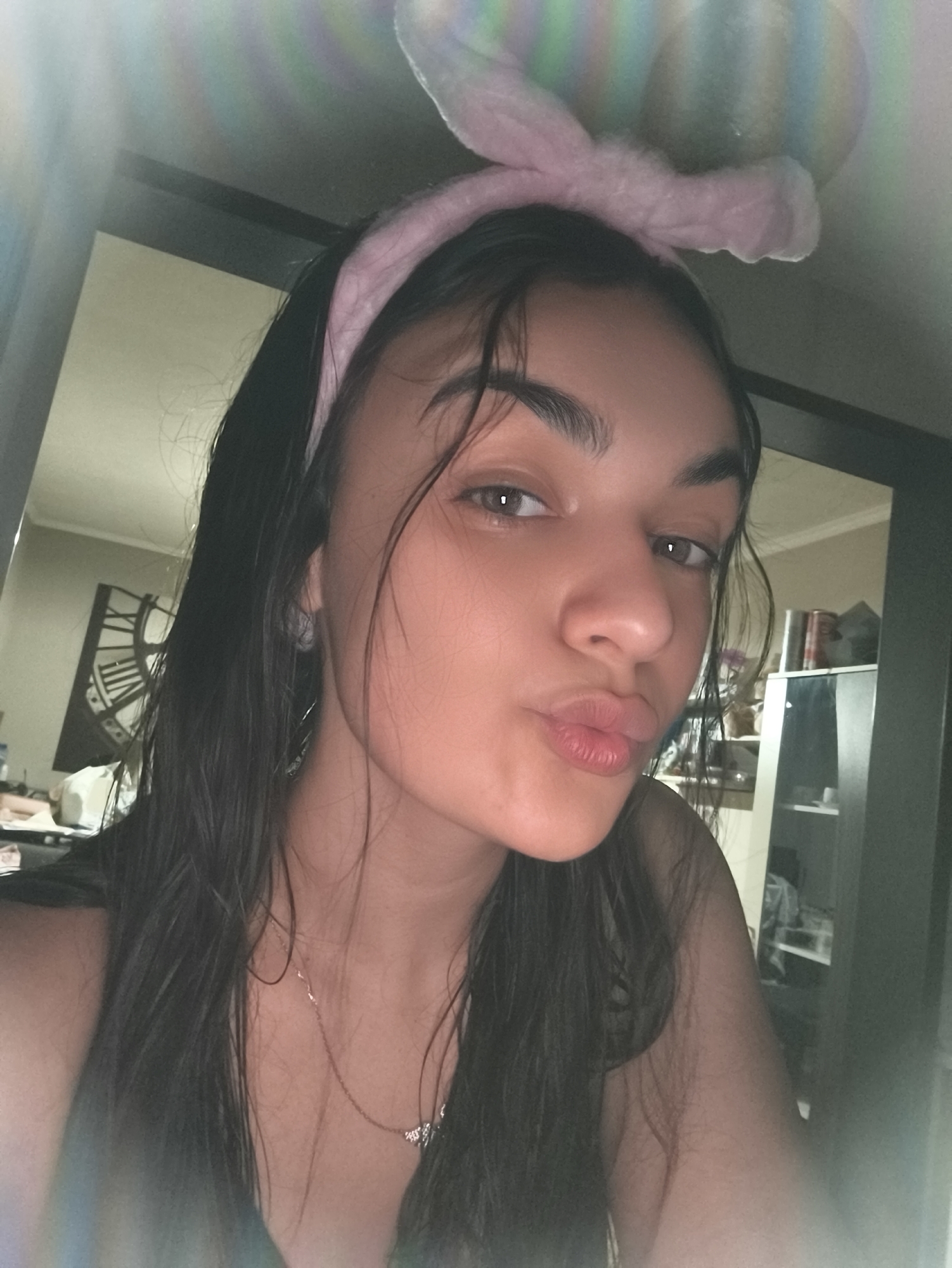 A young woman with dark, wet hair and pink bunny ears on her head poses for a selfie. She is wearing a dark top and a delicate necklace. She puckers her lips for the camera. The background appears to be an indoor setting with furniture and a clock visible on the wall. The lighting is dim and appears to have a lens flare.