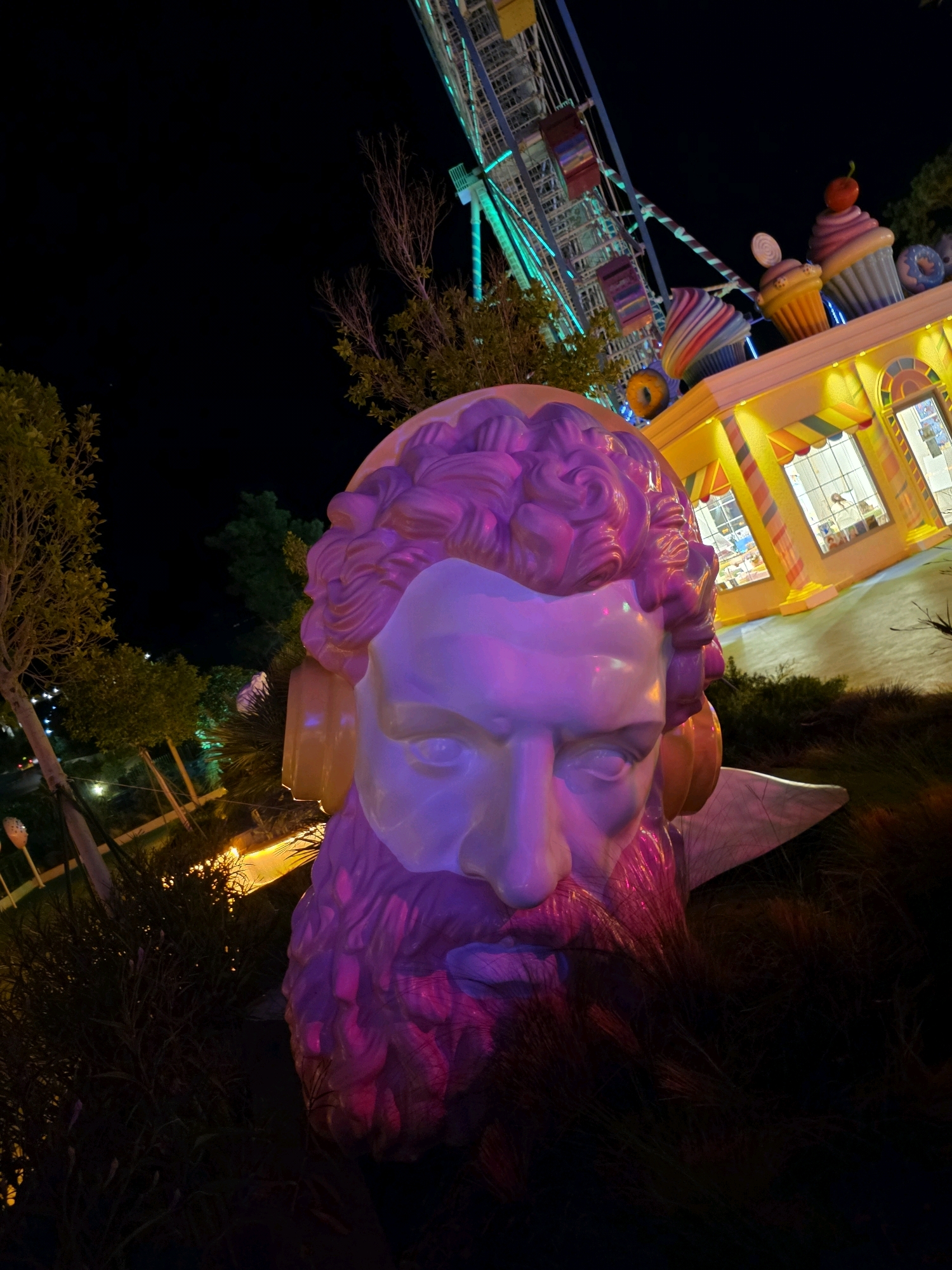 In the city of Bodrum, Turkey, a nighttime scene unfolds at an amusement park, likely a themed attraction, given the whimsical decor. The foreground is dominated by a large, stylized bust of a man's head, rendered in a light purple hue with vibrant pink accents illuminating the beard and hair. The sculpture appears to be made of a smooth, polished material, possibly resin or fiberglass. It is positioned outdoors, amidst dimly lit foliage and sparse landscaping.
In the background, a brightly lit structure resembling a candy shop or food stall stands out with its yellow facade and colorful awnings. Above this building, a Ferris wheel with illuminated cabins, primarily in vibrant green, is visible against the dark night sky. The Ferris wheel appears to be in operation or lit for display. Decorative elements resembling oversized cupcakes and donuts are also present on the roof of the building, further emphasizing the playful, sugary theme of the location. The overall impression is one of a festive and visually striking amusement park environment.