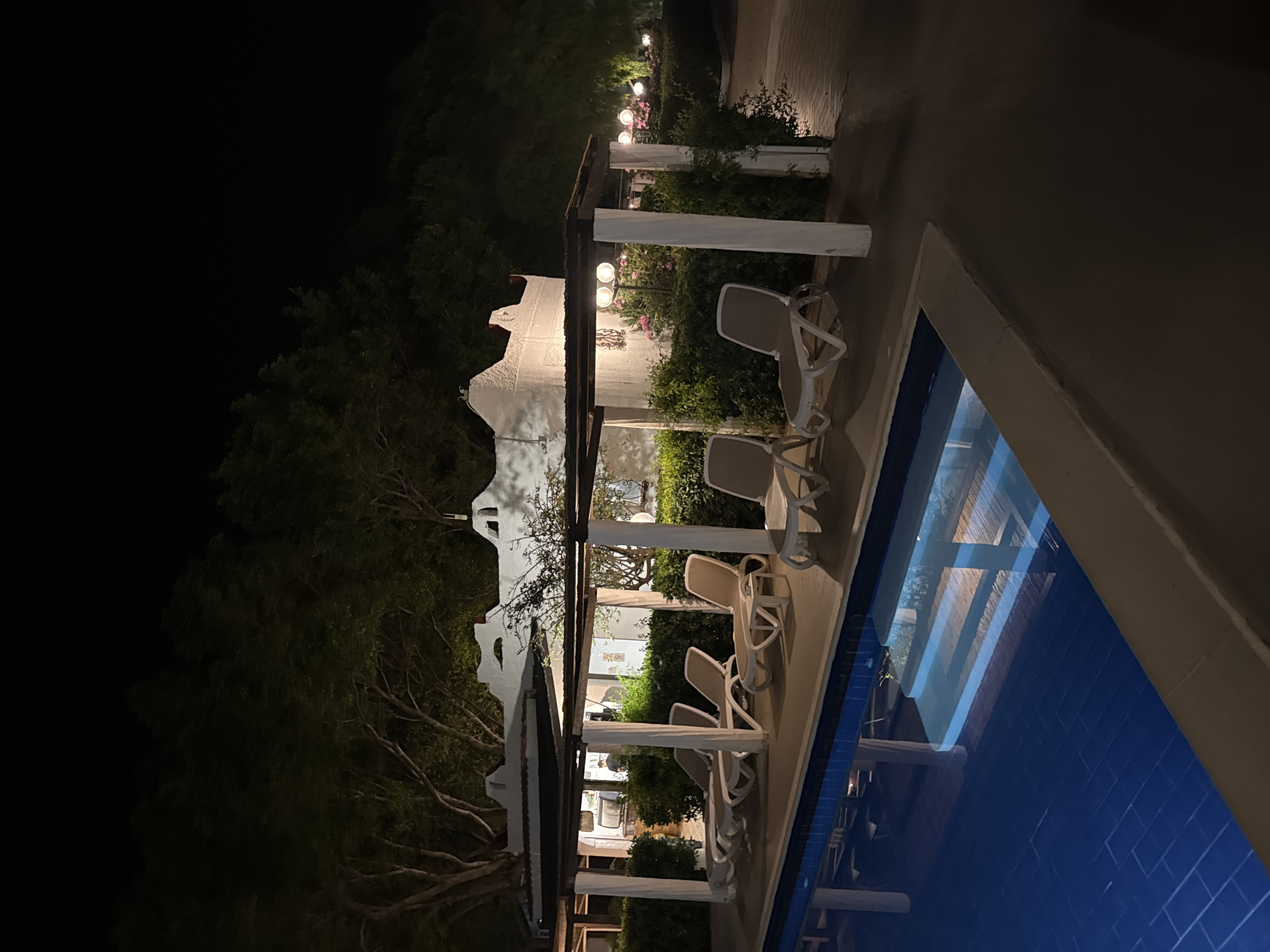 The image depicts an outdoor night scene in Isola delle Femmine, Italy, featuring a swimming pool, lounge area, and illuminated buildings and landscaping. In the lower foreground, a rectangular blue swimming pool is visible, with light-colored steps submerged at the far end and reflecting overhead light sources. Along the near edge of the pool, six light-colored sun loungers are arranged on a paved surface. These loungers are situated beneath a pergola-like structure composed of dark horizontal beams supported by white vertical columns.
Behind the loungers and pergola, a dense line of green hedges separates the pool area from the background. To the left, a white building with distinct architectural features is partially visible; a solitary person is discernable within a dimly lit interior section, appearing to stand behind a counter or bar. To the right, additional sections of the white building are present, alongside a landscaped area featuring multiple spherical outdoor globe lights interspersed among bushes and what appear to be pink flowering plants. Tall, dark trees occupy the upper background, extending into the black night sky, indicating a heavily vegetated environment. The overall lighting is artificial, casting various illuminated zones against the dark surroundings.