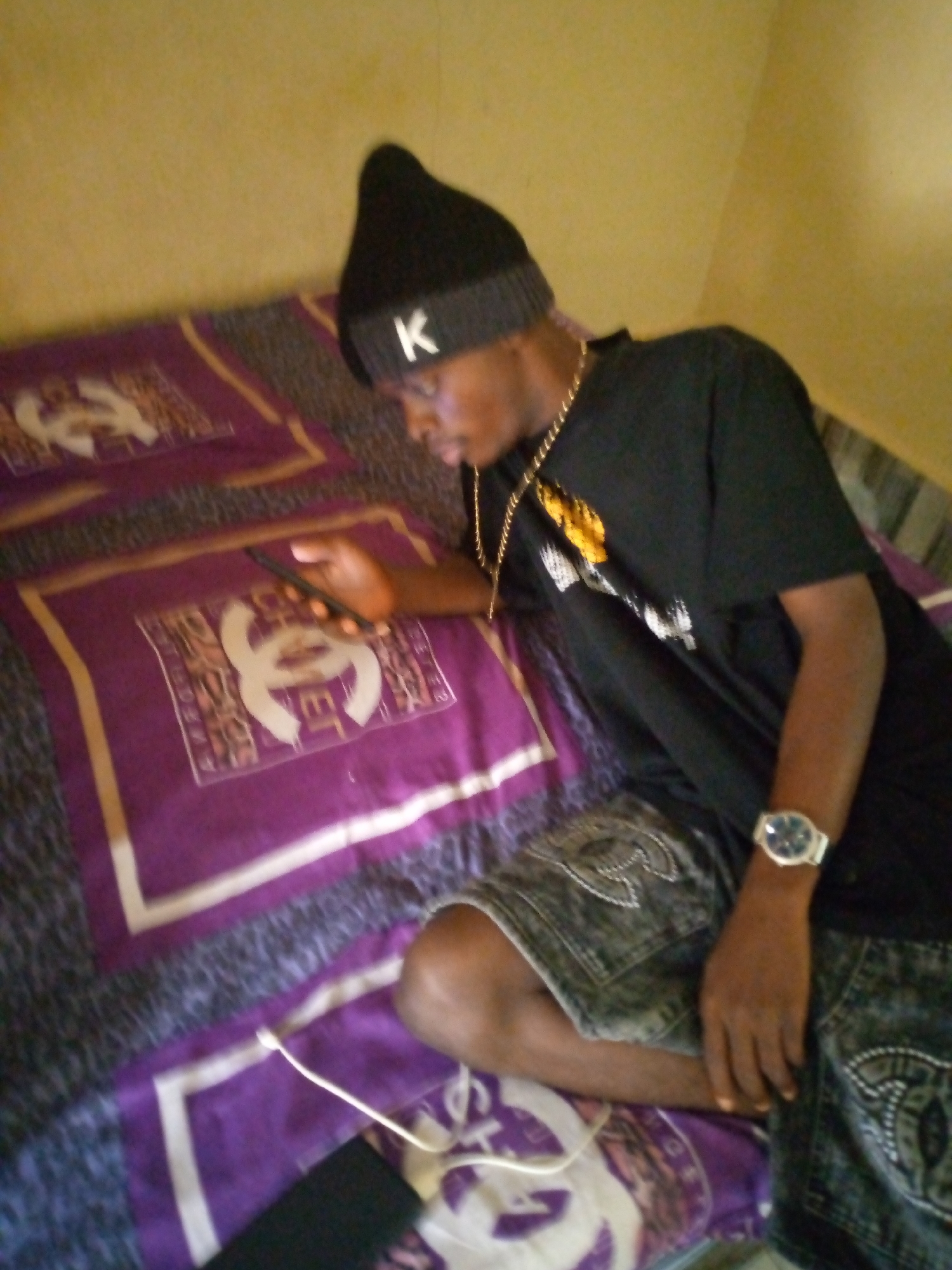A young Black male wearing a black beanie with a white "K" emblem, a black t-shirt with yellow and white graphic print, and distressed denim shorts with a white emblem on the pockets is seated on a bed. He is holding a black mobile phone in his right hand and looking down at it. A gold chain is around his neck, and a white-faced wristwatch is on his left wrist. The bed is covered with a purple blanket featuring a repeating interlocking "C" logo pattern, and a dark grey and white patterned comforter. The background wall is a muted yellow color. The scene appears to be indoors, likely a bedroom in Jalingo, Nigeria.