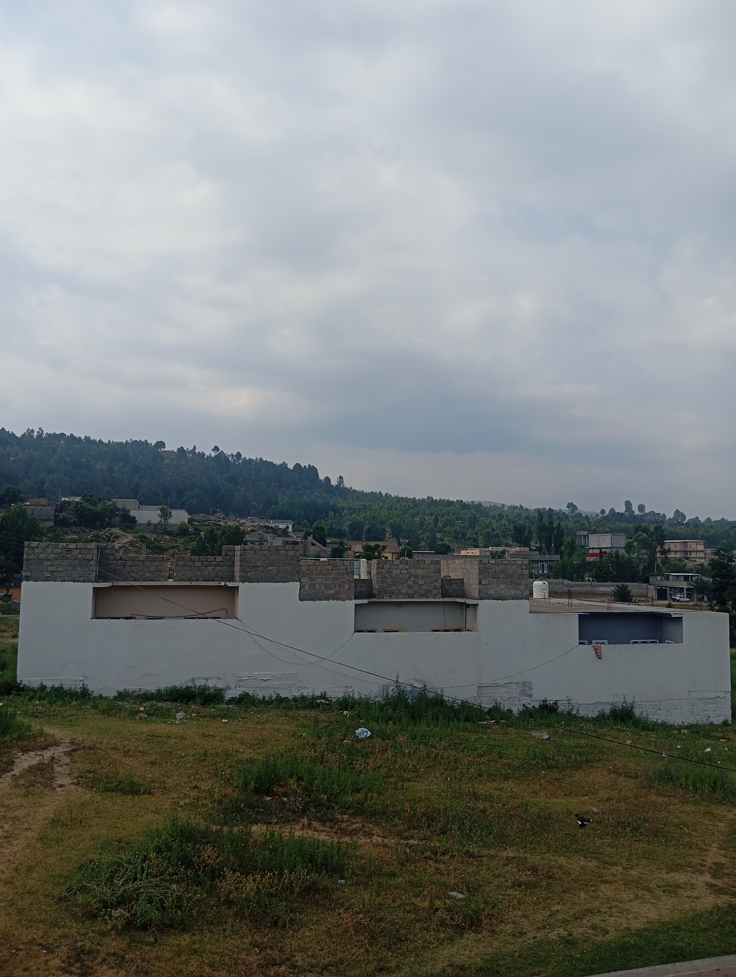 The image depicts a partially constructed building in Lassan, Pakistan. The building is white with visible brickwork on the upper levels. In the foreground, there is a grassy area with some scattered debris. The background features a densely forested hillside dotted with more buildings. The sky is overcast with gray clouds. The scene appears to be an outdoor, daytime view of a semi-urban or rural area. There are no people visible in the image.