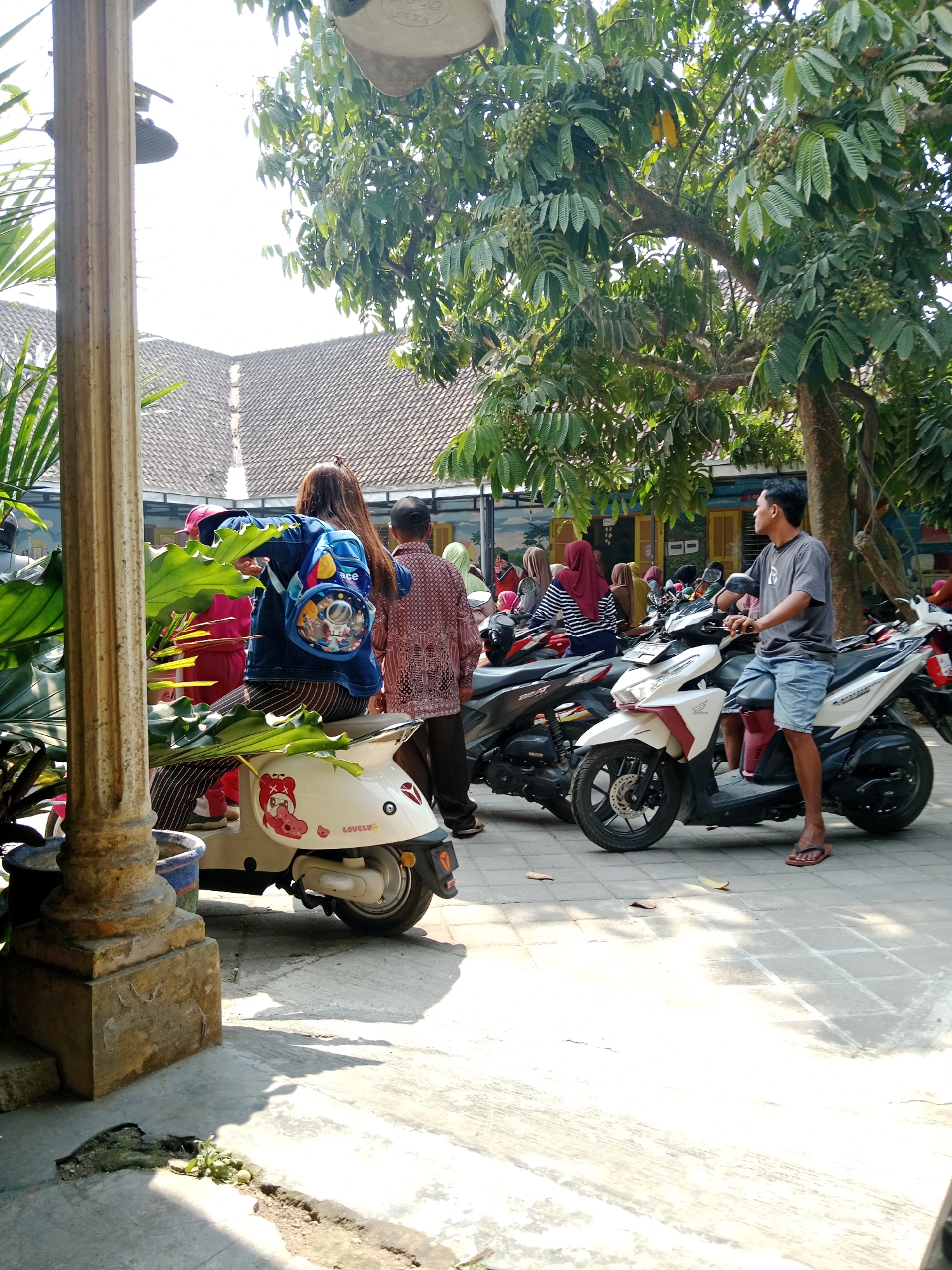 In Srikaton, Indonesia, a street scene unfolds under a bright, sunny sky, with dappled light filtering through the dense leaves of a large tree overhead. A paved walkway, with visible cracks and patches of greenery pushing through, forms the ground. In the foreground, a weathered, fluted column stands to the left. Several motorcycles are parked, forming the central focus of the image.
To the left, a woman sits on a white scooter decorated with a cartoon pig sticker and the word "LOVELY." She wears a denim jacket and has a colorful backpack, featuring cartoon characters, slung over her shoulders. Beside her, partially obscured, is a person in a bright pink outfit.
In the middle ground, another woman, wearing a red headscarf and a striped top, sits on a dark-colored scooter. To her right, a man wearing a grey t-shirt and denim shorts is seated on a white and red scooter, looking off to the right. Several other motorcycles are visible, some occupied by people in various attire, including headscarves and patterned shirts. A man with a patterned shirt and dark pants stands to the right of the white and red scooter. The background shows the distinctive tiled roofs of buildings, typical of Indonesian architecture, and a glimpse of colorful murals on a wall. The overall atmosphere is one of a busy, casual outdoor gathering or a parking area.
