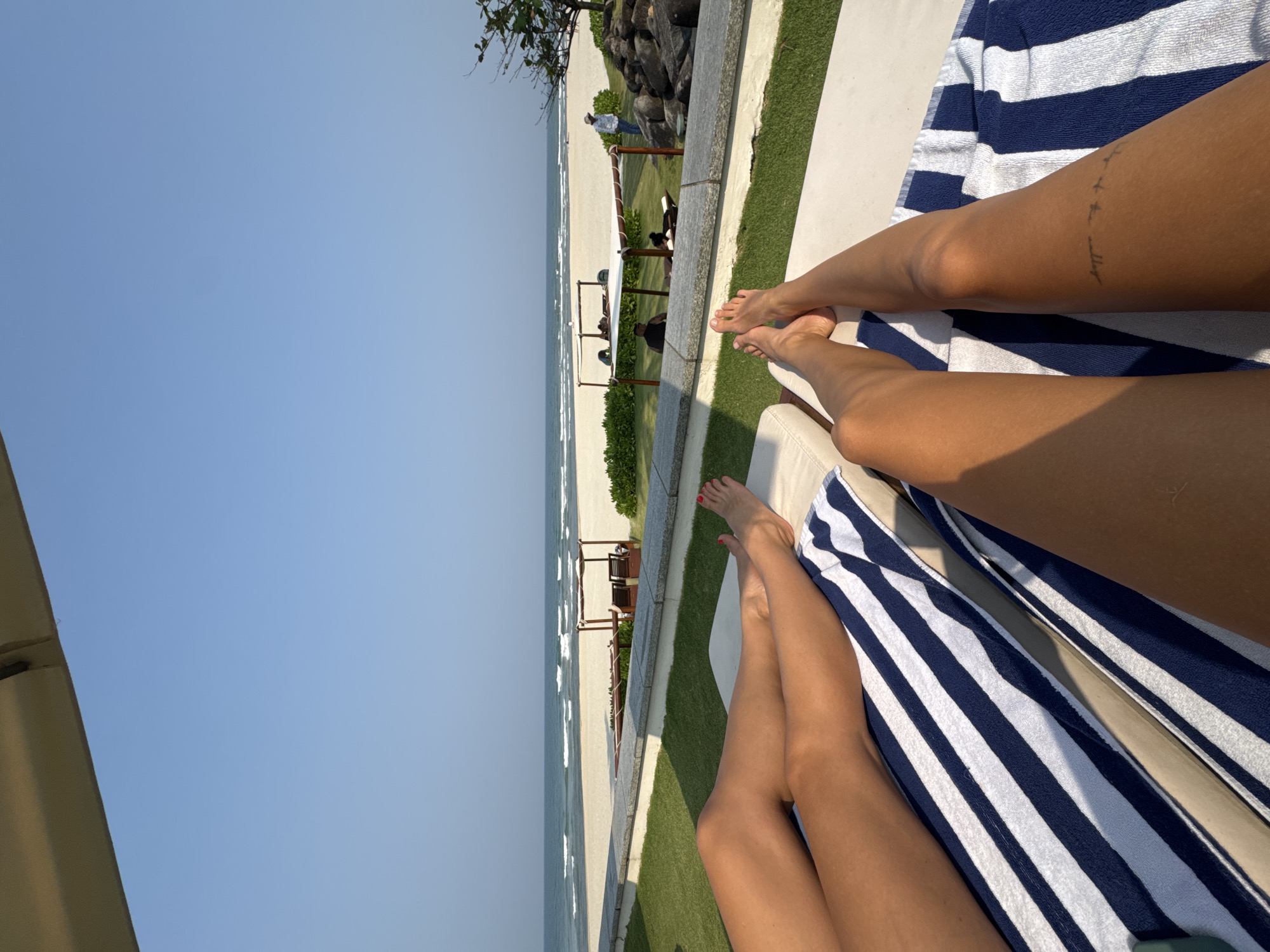 The image shows a view from a beachside lounge chair in Đà Nẵng, Vietnam. Two people are relaxing on adjacent lounge chairs with blue and white striped towels. The scene overlooks a beach with waves breaking on the shore. Beyond the lounge area, a grassy strip separates the seating area from the sandy beach. Several beach umbrellas and chairs are visible in the distance. The sky is clear and blue.