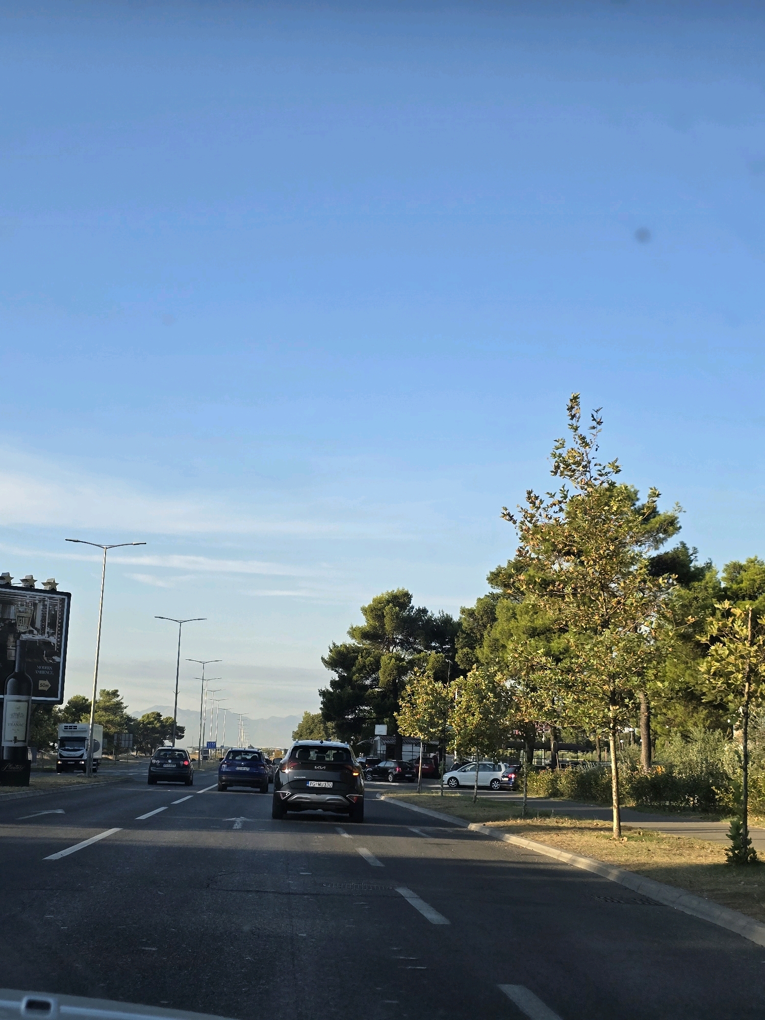 The image depicts a street scene in Podgorica, Montenegro, with cars driving on a multi-lane road. The perspective is from inside a vehicle. On the left side of the road, there is a billboard and a row of streetlights. Beyond the road, trees line the median and the right side of the street. The sky is clear and blue with some wispy clouds. Several cars are visible on the road, including a dark SUV in the foreground, a blue sedan behind it, and a dark hatchback further ahead. A white truck is seen in the distance on the left lane. The road has white lane markings. The environment suggests a sunny day in an urban or suburban area.