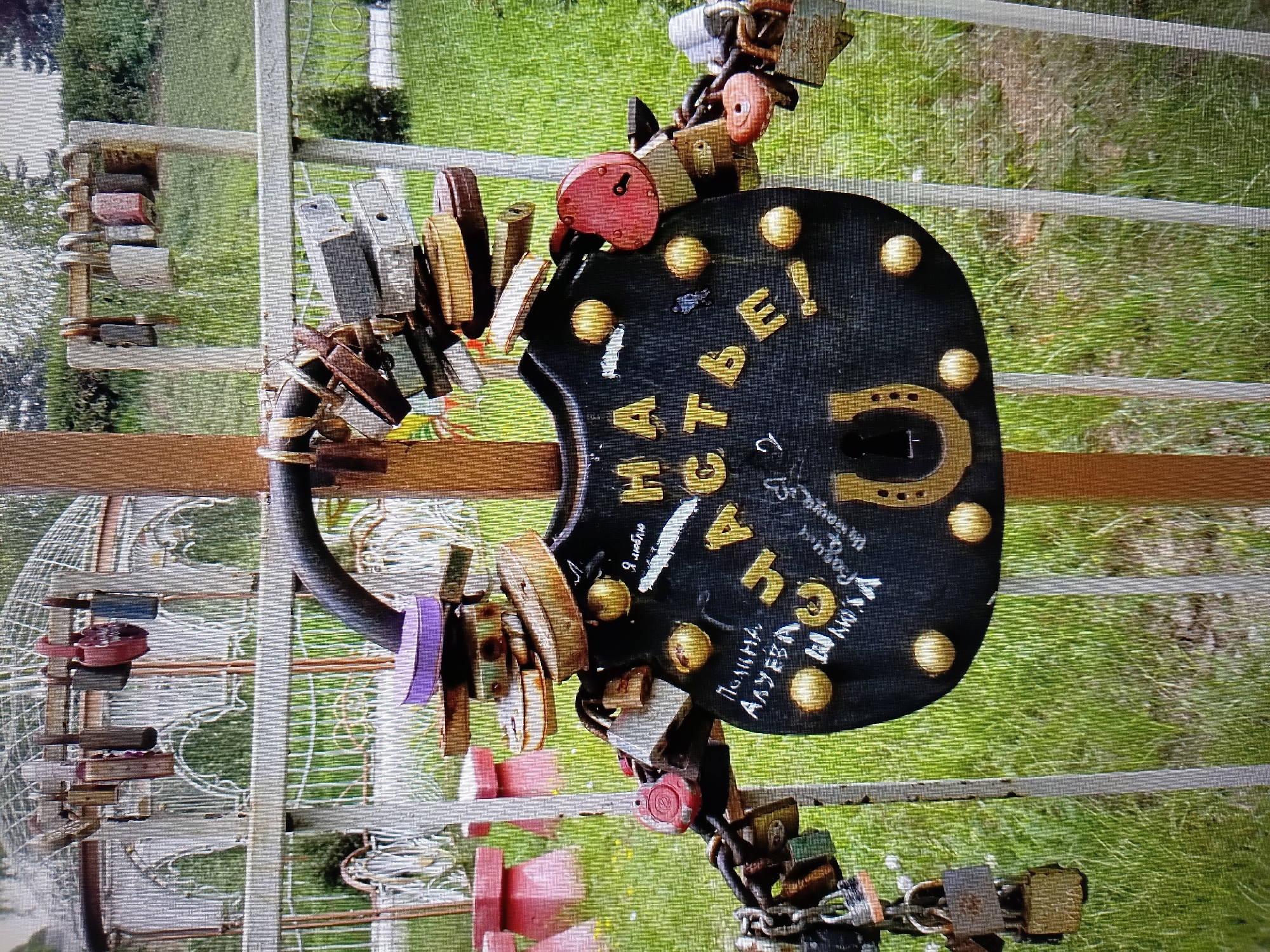 In Tiraspol, Moldova, a black heart-shaped object adorned with gold lettering and a horseshoe is chained to a metal fence. Gold lettering on the object reads "НА СЧАСТЬЕ" which translates to "for happiness". Scattered around the object and chained to the fence are numerous padlocks of various sizes, colors, and states of wear, some with chains still attached. The fence is surrounded by green grass and foliage, with a white gazebo structure visible in the background. Several small pink concrete structures are also present in the background. The overall scene suggests a location associated with good luck charms or romantic gestures.