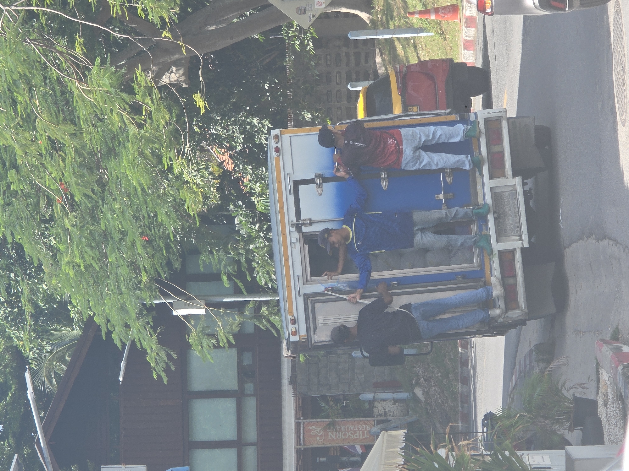 Three men are standing on the back of a white truck with blue doors, in what appears to be a street in an urban environment. The truck is parked or moving slowly on a paved road. The men are dressed in casual clothing. The man on the left is wearing a black t-shirt and blue jeans. The man in the middle is wearing a blue jacket and pants. The man on the right is wearing a red shirt and light-colored pants. The background shows trees, buildings, and a red car. The scene is outdoors, under daylight.