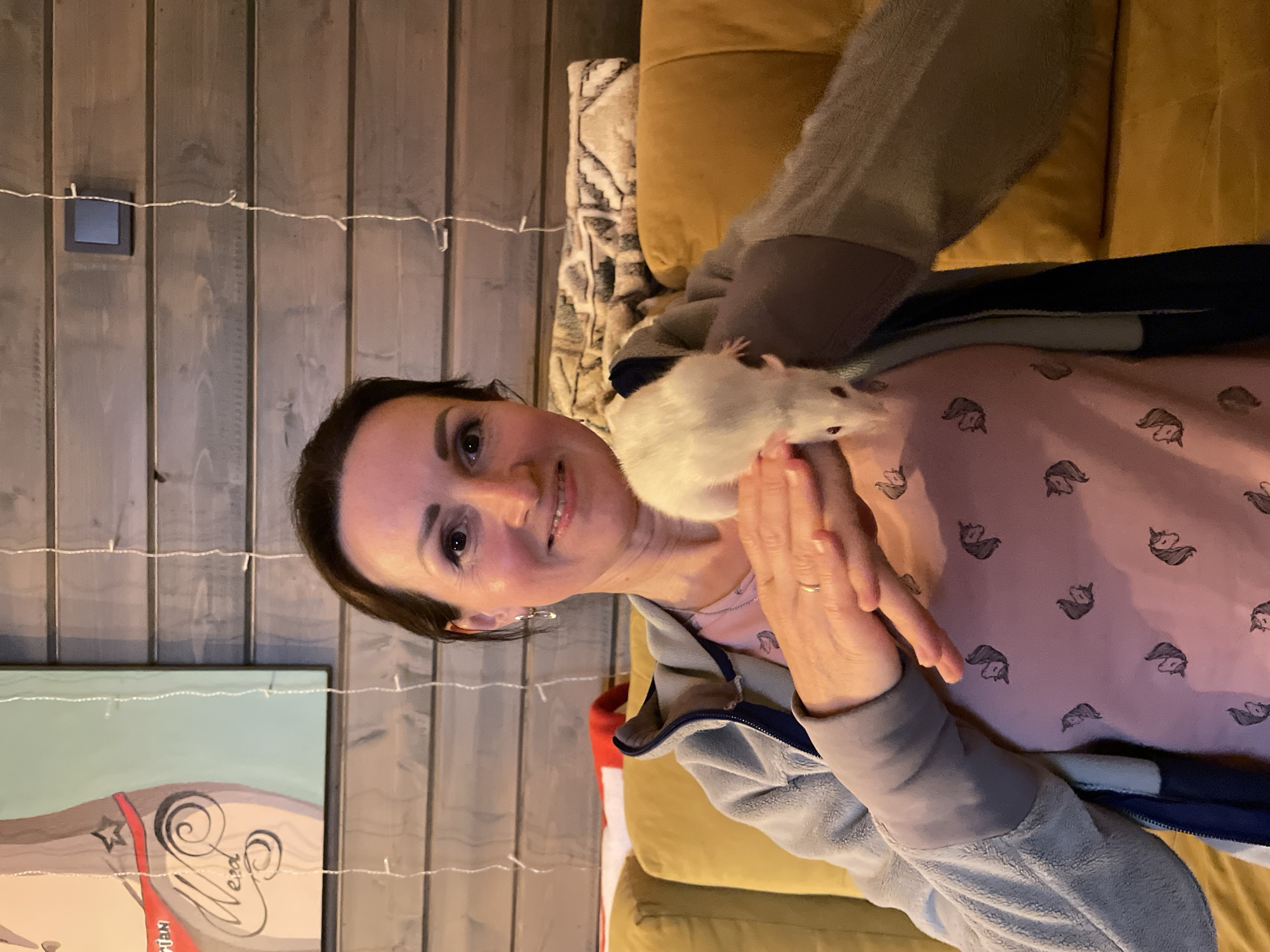 A woman with dark hair and fair skin is pictured from the chest up. She is smiling at the camera while a white and grey rat sits on her shoulder. The woman is wearing a pink shirt with a pattern of unicorn heads and a light blue zip-up jacket. She has a ring on her left ring finger and is holding her hands up. The background consists of grey wood paneling and a painting of a woman with an artistic signature. In the top right corner, there is a portion of a beige sofa and a decorative pillow. The scene appears to be indoors, possibly in a home. The location in the city of Solov'evka, Russia, cannot be independently confirmed from this image.