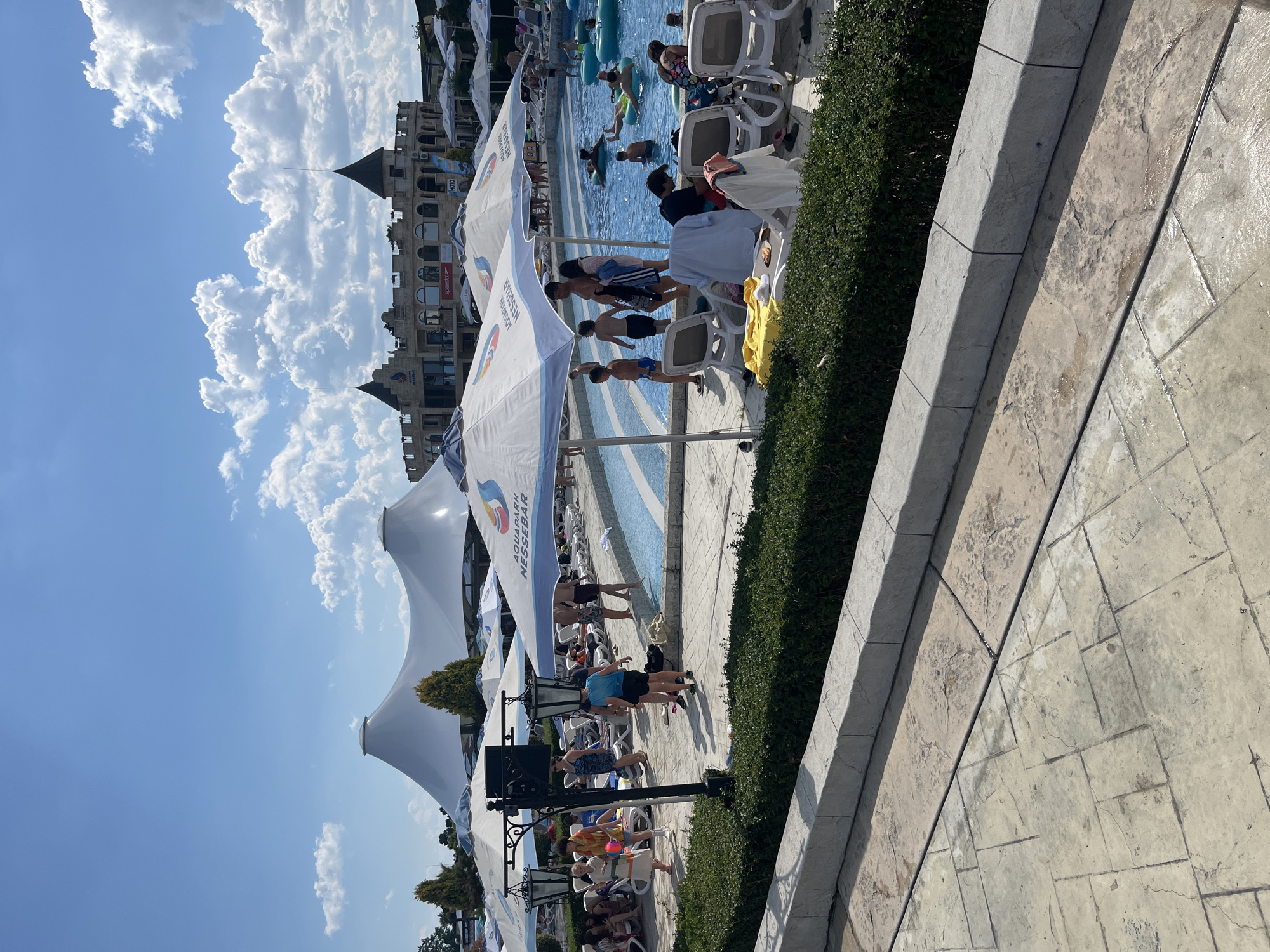 This is a daytime, eye-level shot of a busy aquapark in the city of Ravda, Bulgaria. The aquapark features a large, blue swimming pool filled with people enjoying inflatable tubes and various pool activities. Around the pool, lounge chairs are set up under large, white umbrellas, some of which display the "AQUAPARK NESSEBAR" logo.
In the background, a tan-colored building with castle-like architectural elements, including turrets and arched windows, serves as a prominent structure. White, tent-like structures are visible further in the distance. Green bushes and trees provide landscaping around the paved pathways and seating areas.
People of various ages are present, some swimming in the pool, others relaxing on lounge chairs, and some walking around the perimeter. The sky is a clear blue with scattered white clouds. The ground is paved with textured stone tiles. The overall scene depicts a lively and recreational atmosphere characteristic of a summer day at a water park.