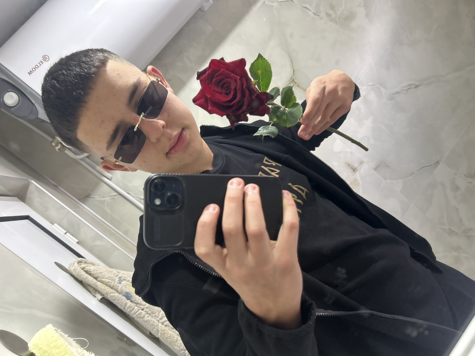 A young person is taking a selfie in a bathroom, holding a red rose in one hand and a black smartphone in the other. The person is wearing sunglasses and a black jacket over a black t-shirt with gold lettering. The bathroom has white marble-patterned tiles and includes a white appliance mounted on the wall. The presence of white towels and bathroom fixtures suggests the scene is a bathroom. No information confirms the scene is specifically located in the city of Zlatograd, Bulgaria, making that aspect speculative.