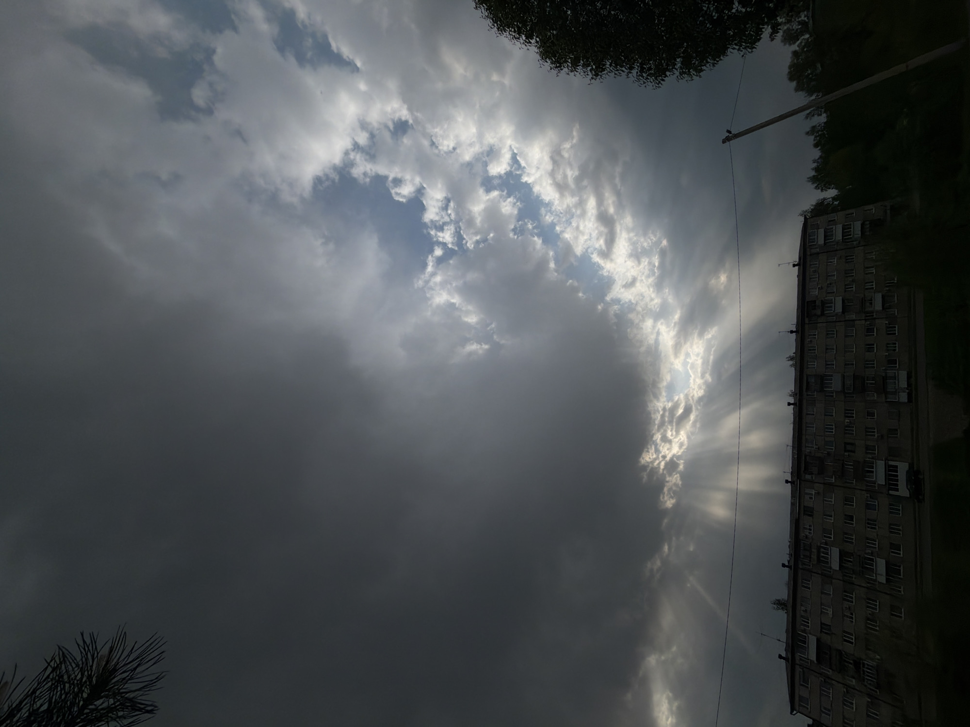 The image depicts a cloudy sky in what appears to be Birobidzhan, Russia, judging by the architectural style of the building on the right. The sky is predominantly covered with dark, heavy clouds, except for a strip of lighter clouds that create a break in the overcast, allowing rays of sunlight to stream through. These rays fall vertically down from the break in the clouds, and slightly illuminate the side of the apartment building, with many windows visible. An electricity pole runs vertically from the upper-center frame down towards the building. Parts of trees are visible from the top and bottom of the image.