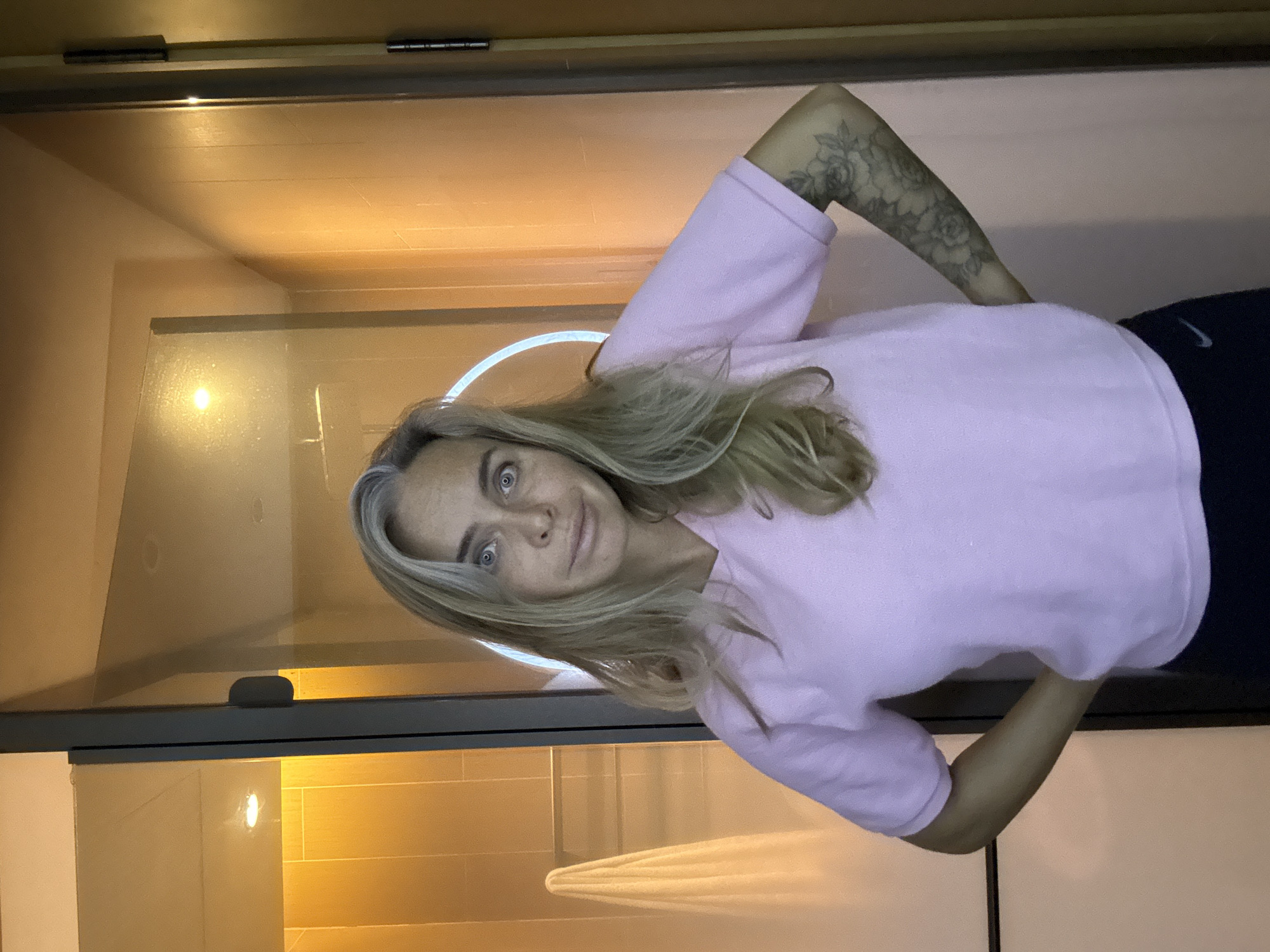 A woman with blonde hair and grey streaks stands with her hands on her hips, looking at the camera with a surprised expression. She is wearing a pink v-neck sweater and dark pants. Her left arm has a floral tattoo. She is standing in what appears to be a bathroom, with a glass shower door and a towel rack visible behind her. The walls are tiled in a warm, orange tone, and the lighting is soft and ambient. There is a circular light fixture visible inside the shower. The overall impression is of a casual, personal moment captured in a hotel bathroom.