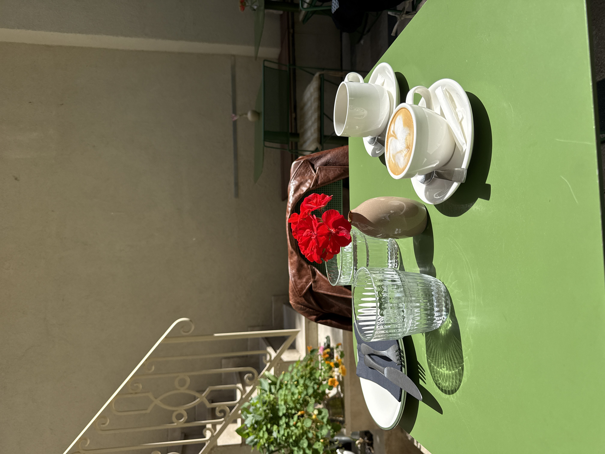The image depicts a still life on a green table in an outdoor setting, likely a cafe or restaurant. Two white coffee cups with latte art sit on saucers, accompanied by spoons. A small vase holds red flowers. Two clear glasses stand near the center of the table, next to a plate with utensils on a dark napkin. A brown leather jacket is draped on a chair partially visible behind the flowers. In the background, there is a white railing of a staircase and a light-colored wall. Green metal chairs and tables are also visible in the background. The scene suggests a sunny day in an urban setting, possibly in Belgrade, Serbia.