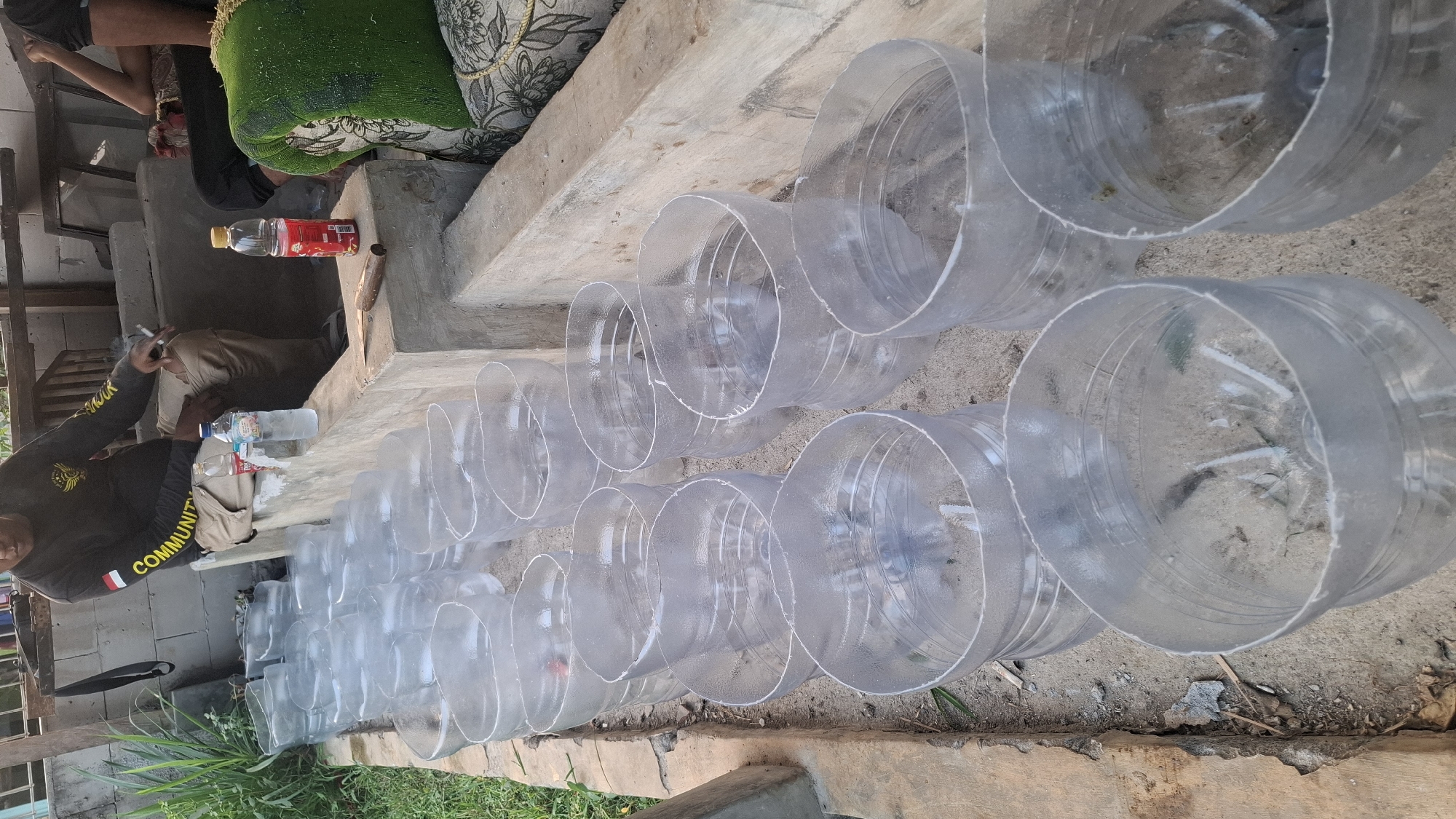 In Rahong, Indonesia, a man is seen sitting on a concrete ledge, smoking. In front of him, a collection of clear plastic bottles, cut to various heights, are arranged in rows on the ground. These bottles appear to be prepared for some purpose, possibly planting or as makeshift containers. Beside the man, on the ledge, are two bottles of water and one bottle of red liquid, suggesting recent consumption or preparation. The background shows a somewhat rustic environment with vegetation and other structures, typical of a residential area in Indonesia.