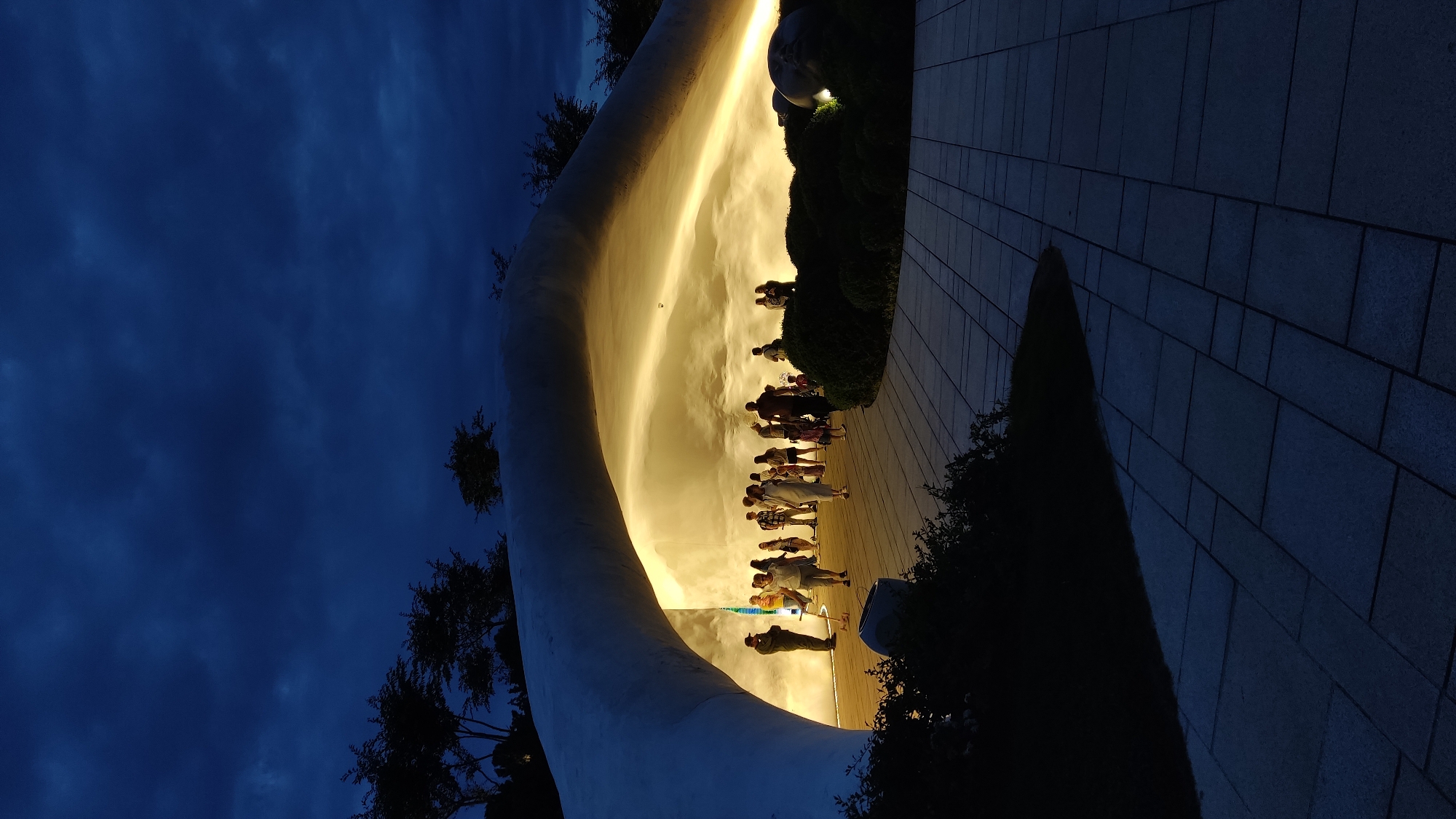 This media file captures an outdoor evening scene in Krasnodar, Russia, likely a public park or plaza, characterized by a striking architectural feature and dramatic lighting. The image, taken at dusk or night, showcases a large, smoothly curved white or light-colored wall or sculpture that dominates the left side of the frame. This structure is brightly illuminated from below with a warm, golden-yellow light, making it glow against the dark surroundings and casting long shadows of people onto its surface.
A group of people, appearing as silhouettes and partially lit figures, are gathered along the base of this illuminated wall, seemingly observing or walking past the installation. Their shapes are clearly visible against the bright background, some standing, others in motion. To the right of the illuminated wall, there's a paved walkway made of rectangular dark tiles, extending into the foreground. Dark, manicured bushes and trees are visible along the right edge and in the mid-ground, further obscuring the background. The sky above is a deep, dark blue, indicating the late hour, with faint patterns of clouds discernible. The overall atmosphere is one of serene contemplation or enjoyment of the illuminated artwork in an urban evening setting. No specific text is visible in the image.