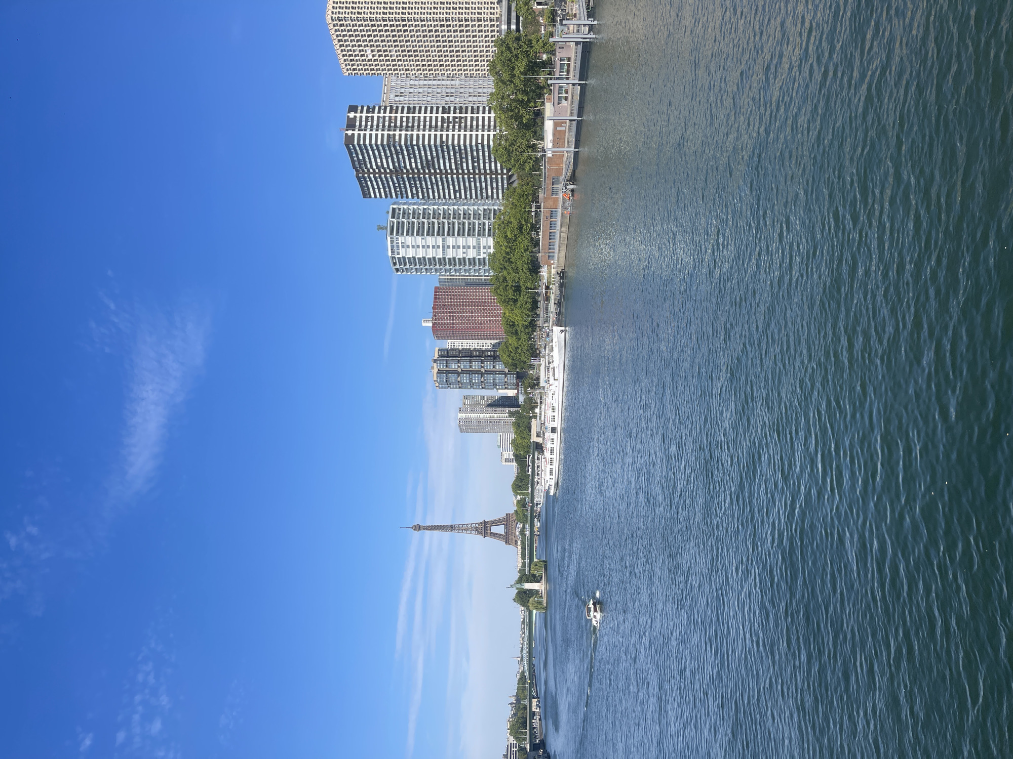 The image captures a scenic view of the Seine River in Paris, France, on a clear day. The Eiffel Tower stands tall in the distance on the left side of the frame, with a small island and a bridge in front of it. A white motorboat is visible in the foreground, moving across the water. Along the right bank of the river, a row of modern high-rise buildings forms the city skyline, interspersed with trees and a waterfront promenade. Several large boats are docked along the bank. The sky is a bright blue with a few wispy clouds.