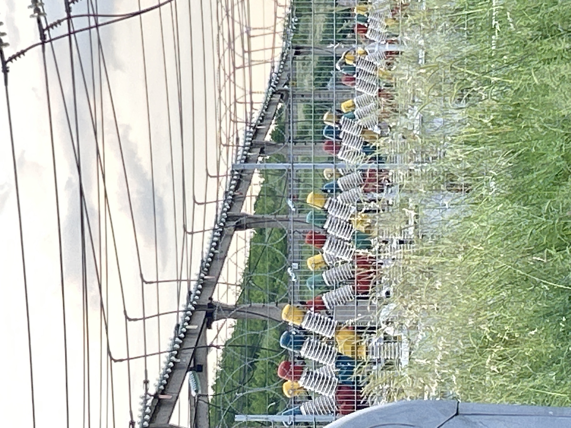 The image depicts a high-voltage electrical substation, possibly on the periphery of Burgas, Bulgaria, judging by the presence of vegetation and infrastructure style typical of Eastern European industrial areas.
Visible elements include:
1. High-voltage transmission lines: Several parallel electrical cables are strung on a support structure, indicating a distribution grid for electrical power.
2. Support structure: A framework made of steel or concrete supports the high-voltage lines, featuring insulators to prevent grounding.
3. Insulators: Series of disc-shaped insulators are attached to the framework to isolate the high-voltage cables. These insulators appear to be multicolored (red, yellow, blue, green).
4. Fencing: A wire fence surrounds the substation equipment, acting as a security barrier. Barbed wire is present on top of the fence.
5. Vegetation: Weeds and tall grass are growing along the fence line, suggesting the area is relatively undisturbed.
6. Sky: The sky is overcast, suggesting the image was taken on a cloudy day.
The overall impression is of a functional but somewhat neglected infrastructure site in an outdoor setting.