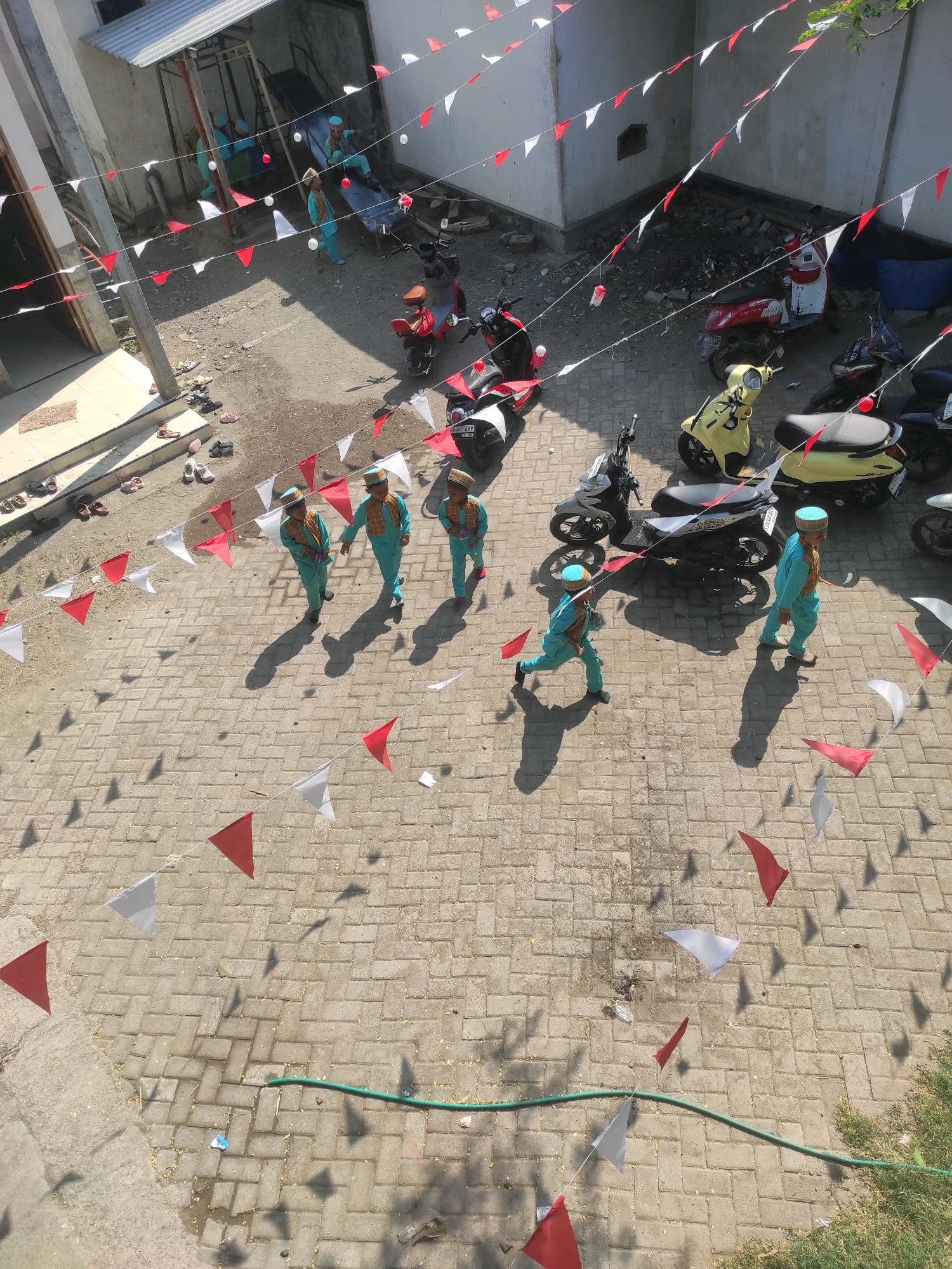 In Klepek, Indonesia, an overhead view captures a paved courtyard decorated with red and white triangular flags strung across. Several children dressed in light blue uniforms and caps are walking or running on the paved surface. In the background, a building with a white facade and a grey roof is visible, with parked motorcycles and a playground structure situated nearby. The scene appears to be outdoors, under bright sunlight, casting distinct shadows from the flags and the people.