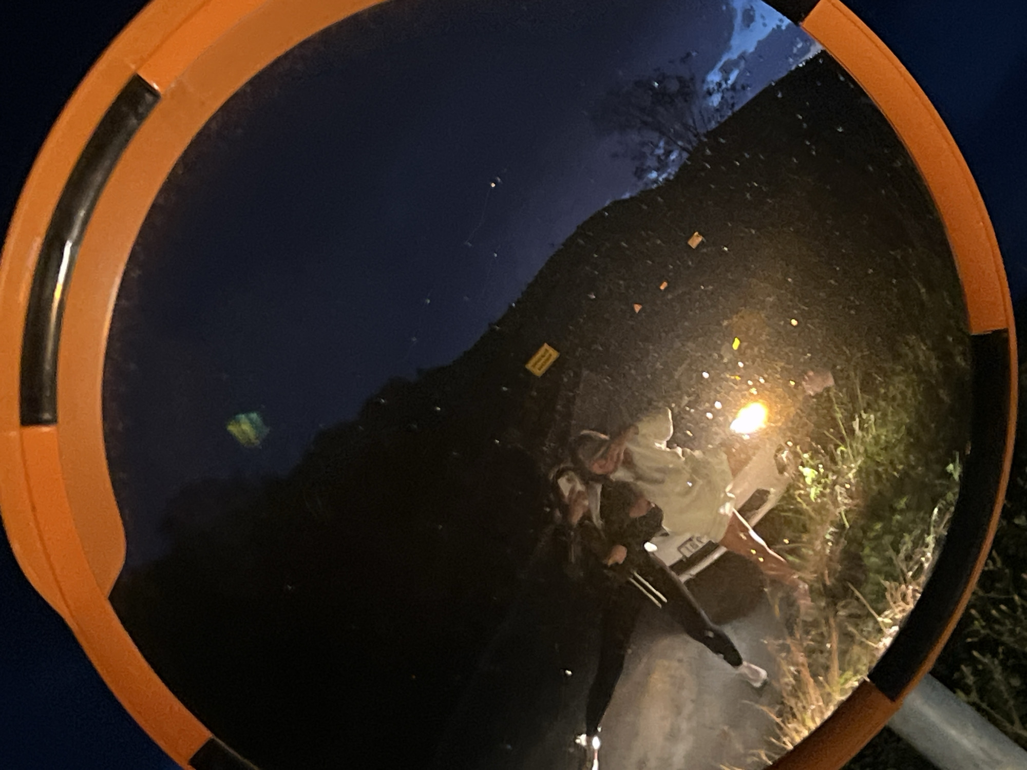 The media file displays a night scene captured through the reflection in a circular convex mirror, likely a traffic or security mirror, framed in orange with black inserts. The overall setting appears to be outdoors at night, indicated by the dark, deep blue sky and the reliance on artificial light sources.
In the reflection, two individuals are visible near a white vehicle, which could be a car or a small van. The vehicle's headlights are brightly illuminated, casting a strong glare that highlights portions of the scene and scatters numerous small, bright specks across the mirror's surface, possibly reflections from dust or moisture. One person, wearing a light-colored top and dark pants with white sneakers, is partially visible near the front of the vehicle, appearing to lean or reach down towards the ground or the vehicle's side. The other person is less distinct, partially obscured behind the first, suggesting they are standing close by.
Lush green and brown vegetation, possibly grass and bushes, lines the side of what appears to be a road or unpaved area next to the vehicle. The distorted reflection also shows a silhouette of trees or distant landscape against the dark sky in the upper right quadrant. A small, rectangular object, too blurry to read, lies on the reflected ground towards the center-left. No other discernible text is visible on the vehicle or elsewhere. The scene is set in Visnjeva, Montenegro, suggesting this is a local road or area at night. The activity appears to be an interaction with or near the parked vehicle under the artificial light.