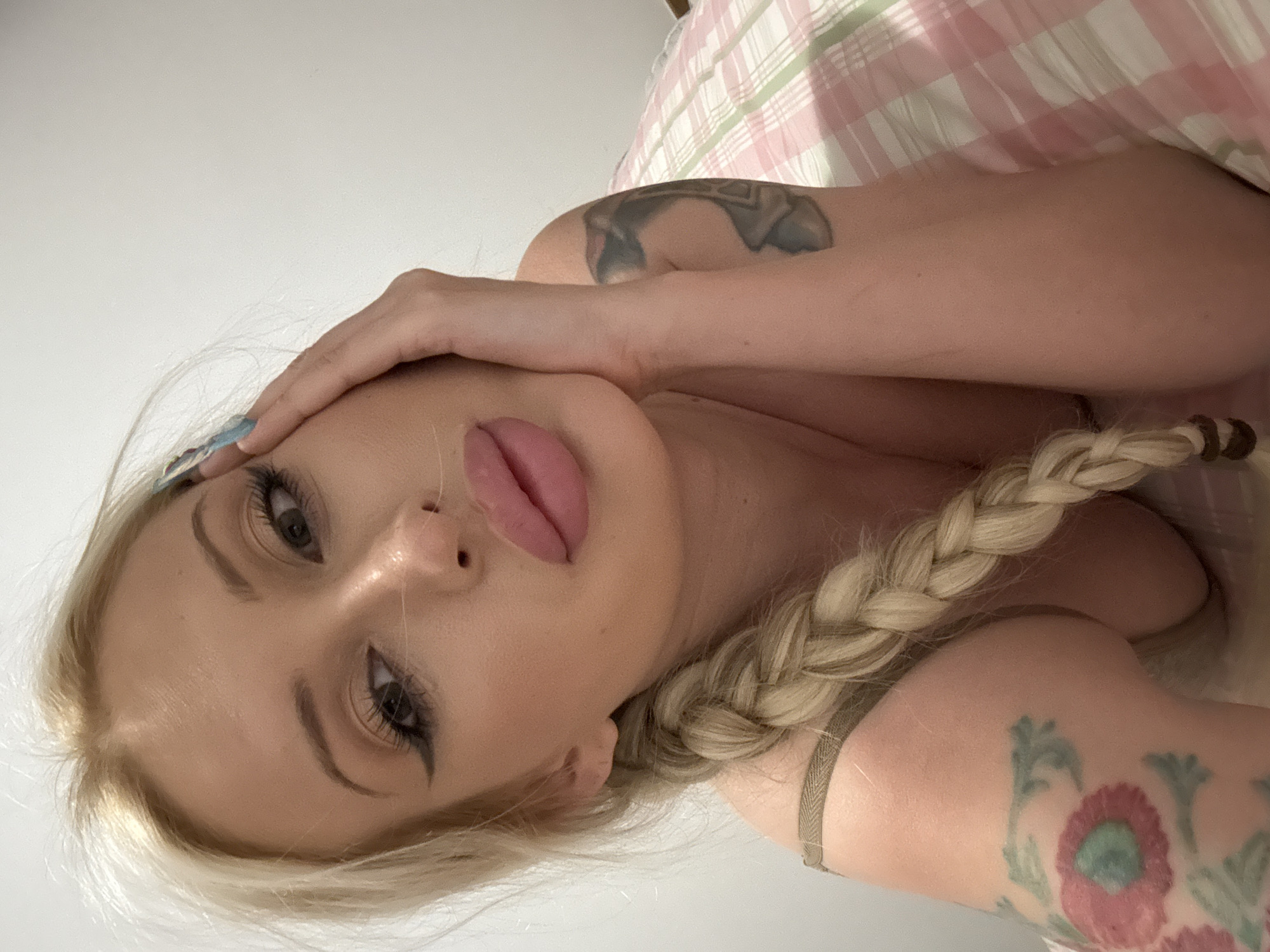 A young Caucasian woman with blonde hair in a braid is lying on her side, propped up on her elbow on a bed. Her right hand is touching her face. She is wearing a tan bra. She has tattoos on her arms. The bed is covered with a pink and white checkered sheet. The background is a plain white wall. The lighting is natural.