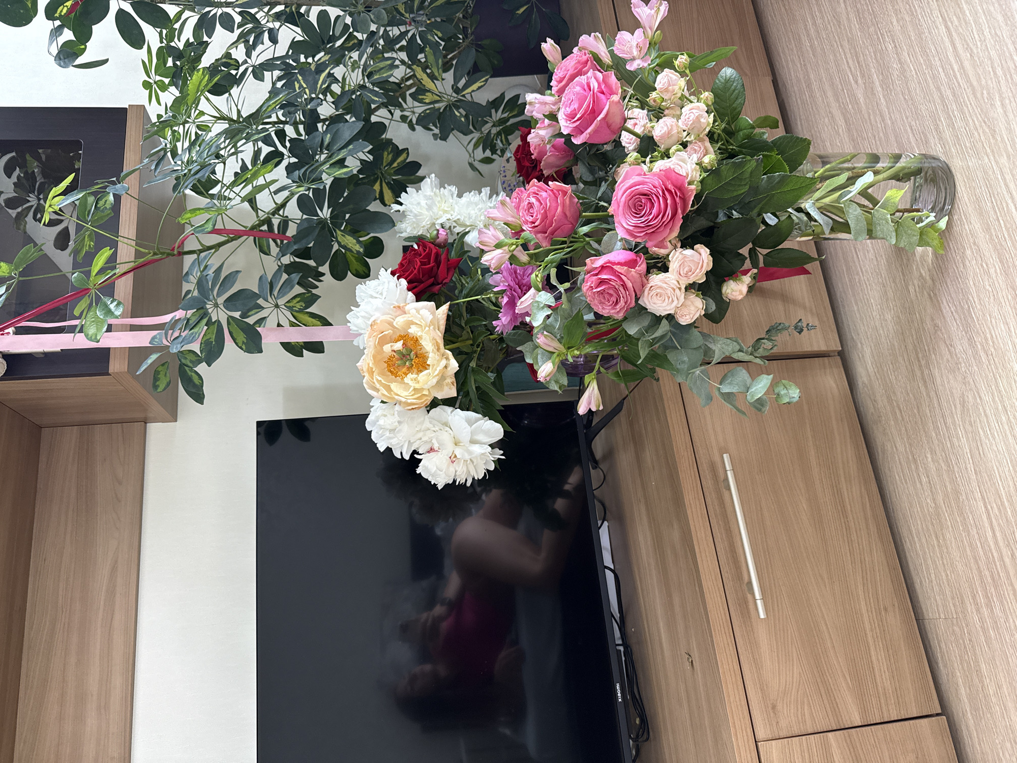 In an indoor setting, a modern entertainment center with wooden drawers and a black television screen is visible. The television displays a reflection of a person lying down, wearing a pink outfit. In front of the entertainment center, a large vase filled with a vibrant bouquet of pink and white roses, along with other flowers and greenery, sits on the floor. Behind the bouquet, a large potted plant with green leaves is situated to the right of the television. The room appears to be a living area, possibly in an apartment, with a wooden floor. There is no indication of the city or country.