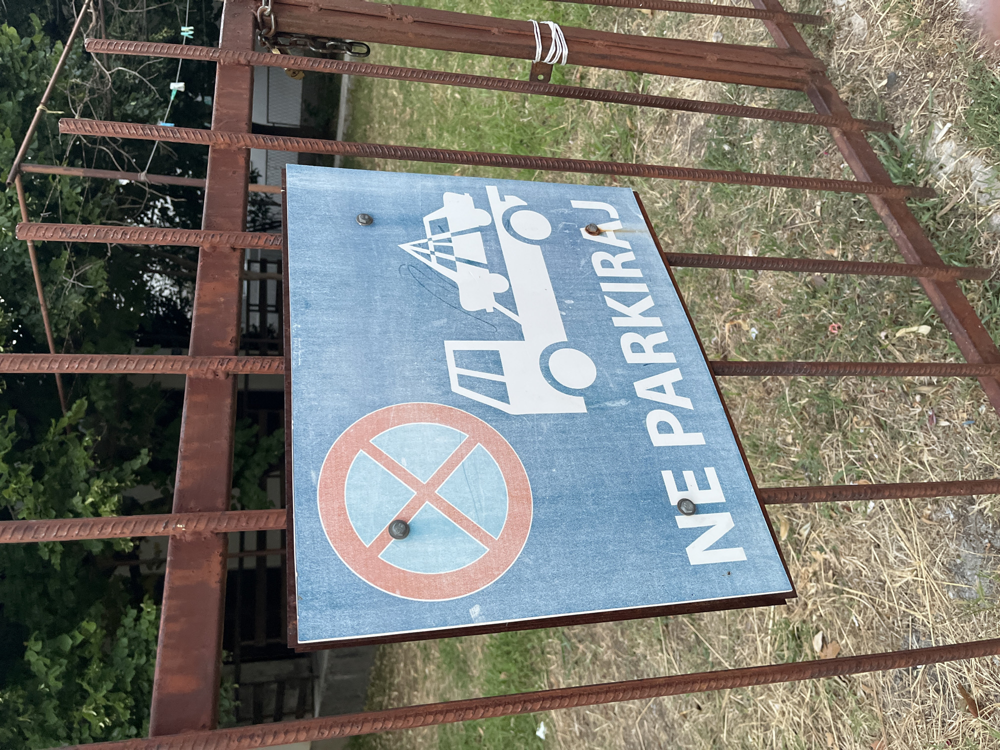 A weathered blue sign reading "NE PARKIRAJ" (Do Not Park) in white letters is attached to a rusty metal fence. The sign also features a graphic of a tow truck lifting a car, indicating that vehicles will be towed if parked here. The fence is made of vertical rebar-like metal bars, some of which are rusted and appear to be part of a gate. In the background, lush green trees and a glimpse of a building are visible. The ground is covered with dry grass and some scattered debris. The sign is secured to the fence with two visible bolts. The overall impression is that of a functional, albeit somewhat neglected, prohibition sign in an outdoor urban or semi-urban setting.