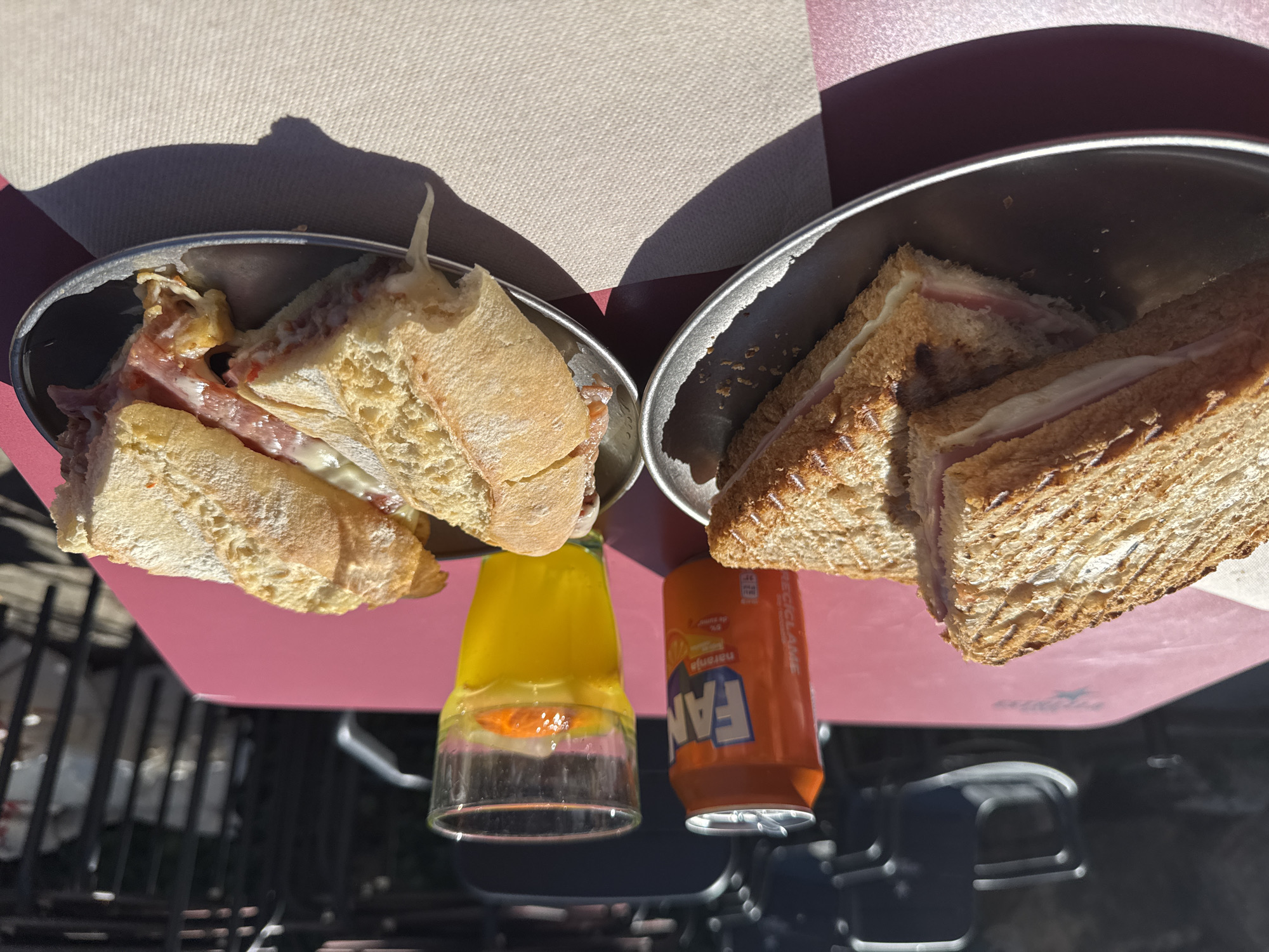 The image shows a table in Alp, Spain, set with two metal bowls containing sandwiches, a glass with orange slices and liquid, and a can of Fanta orange soda. The sandwiches in the left bowl are made with crusty bread, ham, and melted cheese. The sandwiches in the right bowl are made with toasted bread, ham, and cheese. The table has a pink and beige surface. The ground below the table is visible through the gaps in the metal frame and shows a dark, indistinct background. The scene appears to be outdoors, given the presence of shadows.