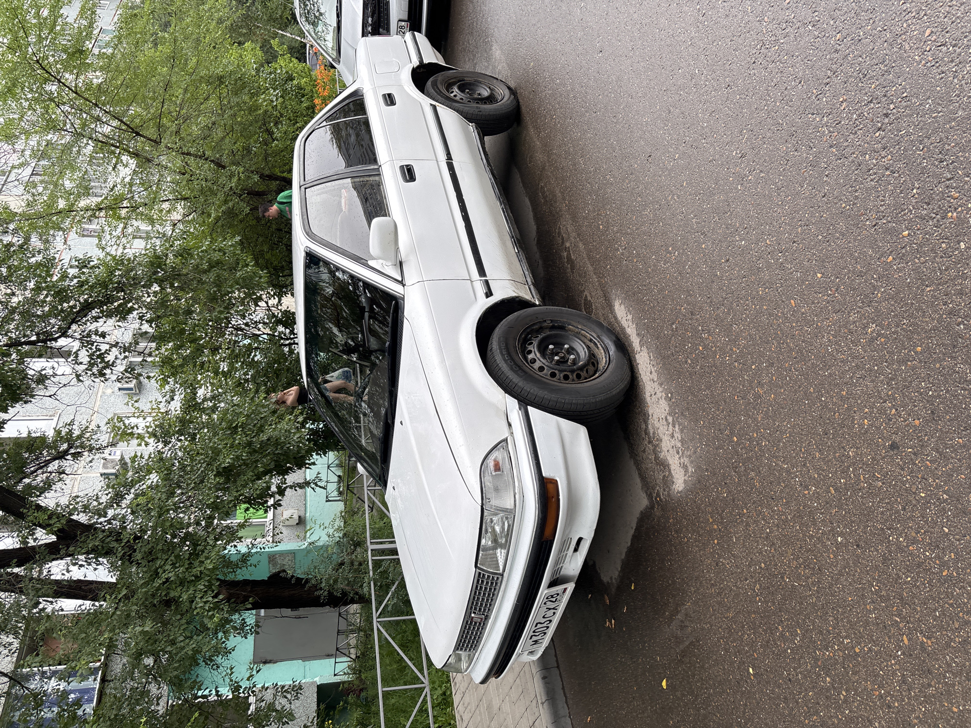 A white sedan is parked on a wet asphalt street next to a sidewalk. The car has a license plate that reads "M303CX 28". In the background, there is a multi-story apartment building with trees and foliage. Two people are visible in the background, one standing near the building entrance and the other walking between trees. The sky appears overcast. The overall scene depicts a typical urban environment in Blagoveshchensk, Russia, possibly on a rainy day given the wet street and pavement.