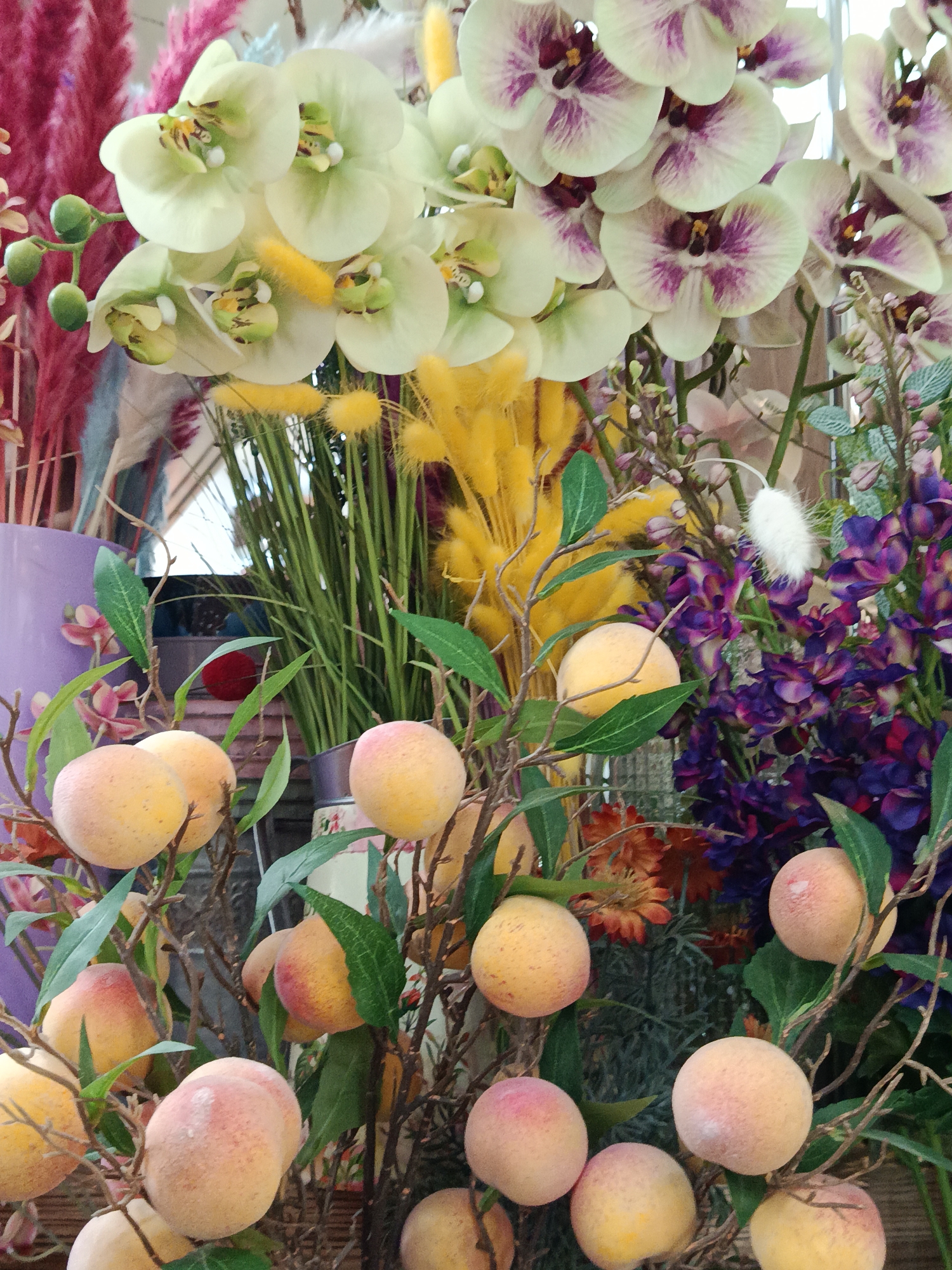The image shows a close-up view of an assortment of artificial floral and fruit arrangements. Dominating the top of the image are two clusters of orchid blossoms, one with pale green petals and yellow centers and the other with white petals and purple markings. Behind these are plumes of yellow and purple flowers. In the foreground are several artificial peaches, attached to brown branches with green leaves. The peaches are a pale yellow color with slight pink hues. The background appears to be a retail display with a variety of colorful artificial plants in vases. The context of the photo doesn't provide information to confirm if this image was taken in Moscow, Russia.