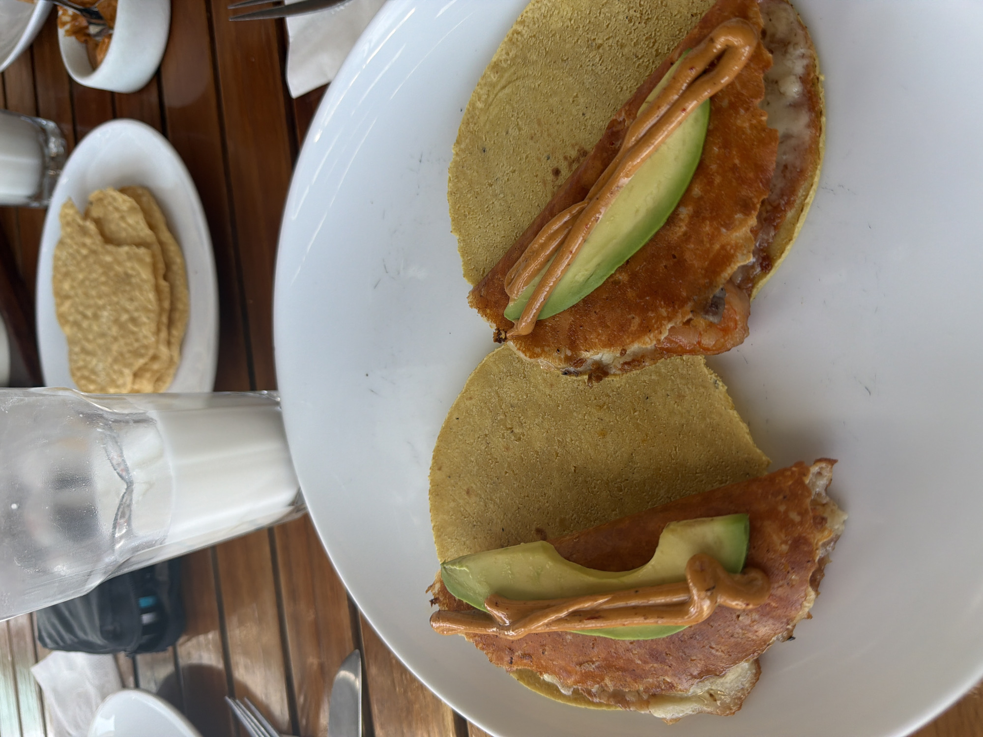 The image captures a close-up, overhead view of a meal served on a wooden table, likely in Puebla, Mexico. The central focus is a large white oval plate holding two yellow, round tortillas. Each tortilla is topped with a generous layer of what appears to be pan-fried or grilled meat, possibly ham or a similar cut. Sliced avocado and a zig-zag drizzle of an orange-hued sauce, possibly a spicy or creamy dressing, adorn the top of the meat.
To the left, a small white oval plate contains several flat, golden-brown, irregular-shaped cracker-like items. Further up and to the left, a small white bowl holds what looks like smaller, fried tortilla pieces or crispy snacks. A clear glass bottle or pitcher filled with white liquid, resembling milk, is positioned towards the bottom-left of the main plate. Utensils, including a fork and knife, are visible on the wooden table, suggesting a full place setting. The warm brown tones of the wooden table contrast with the white crockery and the vibrant colors of the food. The lighting suggests daytime, and the overall scene depicts a casual dining experience. There is no visible text in the image.