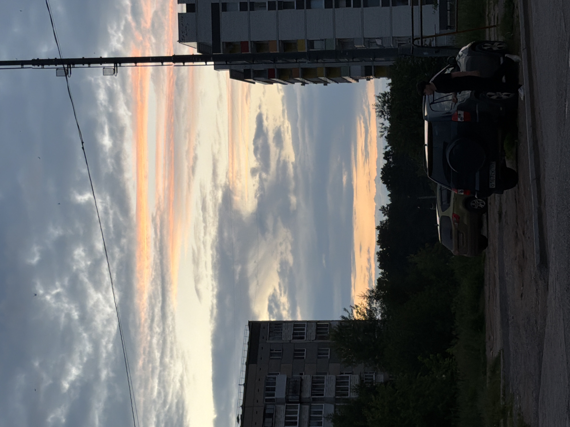 The image depicts an outdoor scene at dusk, likely in Chaikovsky, Russia, with a striking sunset sky featuring bands of pale orange and pink clouds against a backdrop of grey. In the foreground, a paved area leads to a dirt and gravel patch. Two cars are parked: a dark-colored SUV with a spare tire on the rear door and a lighter-colored hatchback to its left. A young man wearing a black t-shirt and black pants, with white sneakers, is walking towards the dark SUV from the right side of the frame.
Dominating the background are two multi-story apartment buildings. The building on the left is older, constructed of grey concrete blocks with numerous balconies and windows. The building on the right is taller, also made of concrete, with colorful accents on some of the balconies. Trees and dense foliage line the space between the buildings and the foreground. A tall utility pole with a ladder and what appear to be fixtures is positioned to the right of the apartment building. The overall impression is of a residential urban environment during the twilight hours.