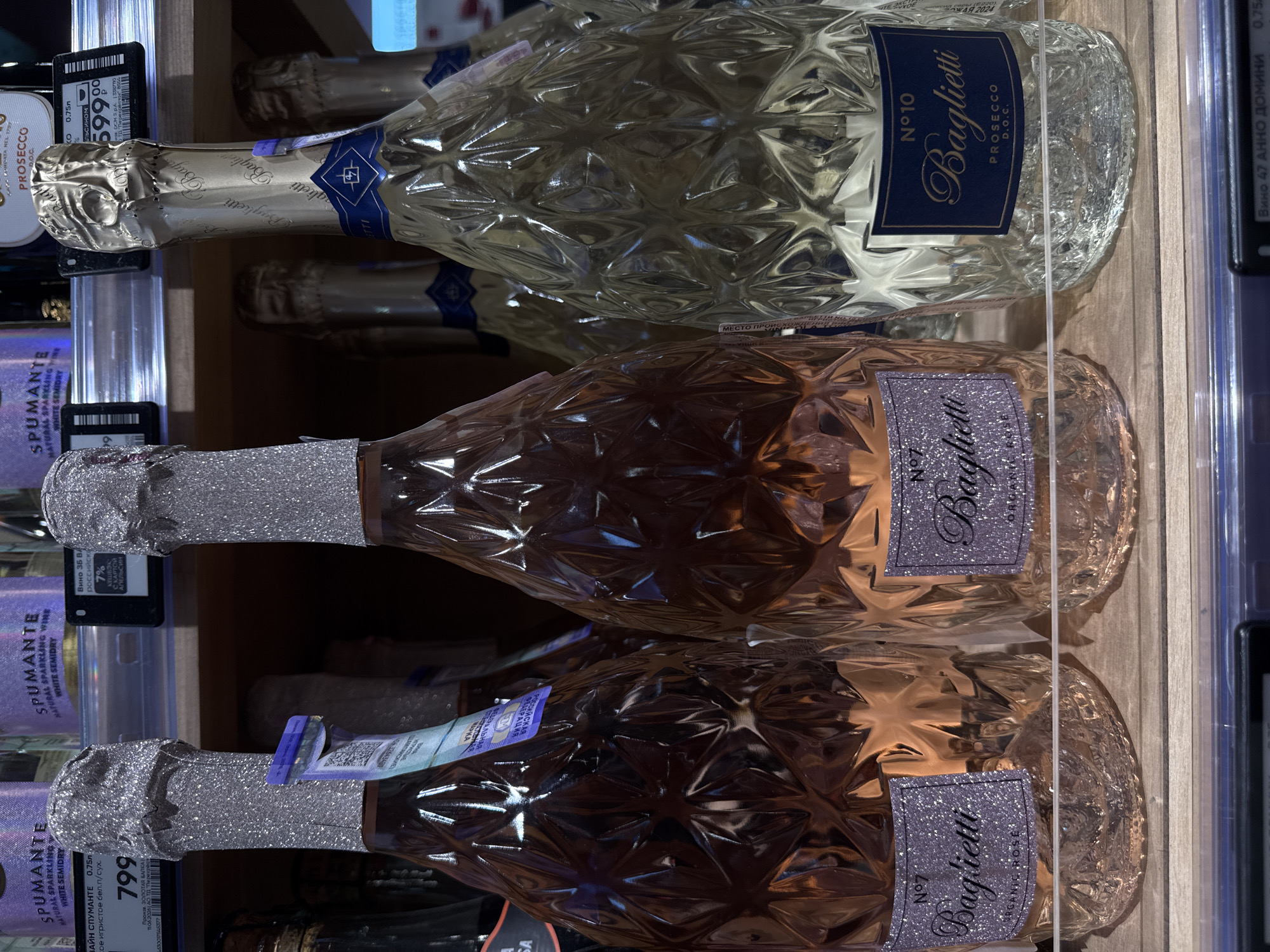 The image shows a close-up, eye-level view of three bottles of sparkling wine displayed on a shelf in what appears to be a retail environment, possibly a grocery store or wine shop, in Moscow, Russia. The bottles are positioned prominently in the foreground, with their distinctive faceted glass bodies and decorative foil-wrapped necks.
The bottle on the far left is a rosé sparkling wine, identified by its pinkish hue and a label that reads "Baglietti N°7 ORGANIC ROSE." The middle bottle is also a rosé sparkling wine, with a similar faceted design and "Baglietti N°7 ORGANIC ROSE" label. To the right is a bottle of white sparkling wine, also with a faceted glass body, and a label that states "Baglietti N°10 PROSECCO D.O.C." The foil on the necks of the rosé bottles is silver and glittery, while the foil on the white wine bottle is a pale gold.
Behind these bottles, a row of additional bottles of sparkling wine is visible, suggesting a larger display. The shelf itself is made of wood. Above the bottles, pricing tags are visible. The tag on the left, associated with one of the rosé bottles, displays a price of "799". Another tag further to the right, associated with the white wine, shows "599". These price tags indicate that this is a commercial setting where products are being offered for sale. The overall lighting is bright and even, typical of retail store lighting. There are no people or animals present in the frame. The context suggests a standard product display in a Russian retail store, potentially in Moscow given the pricing units and Cyrillic text on some of the labels or price tags.