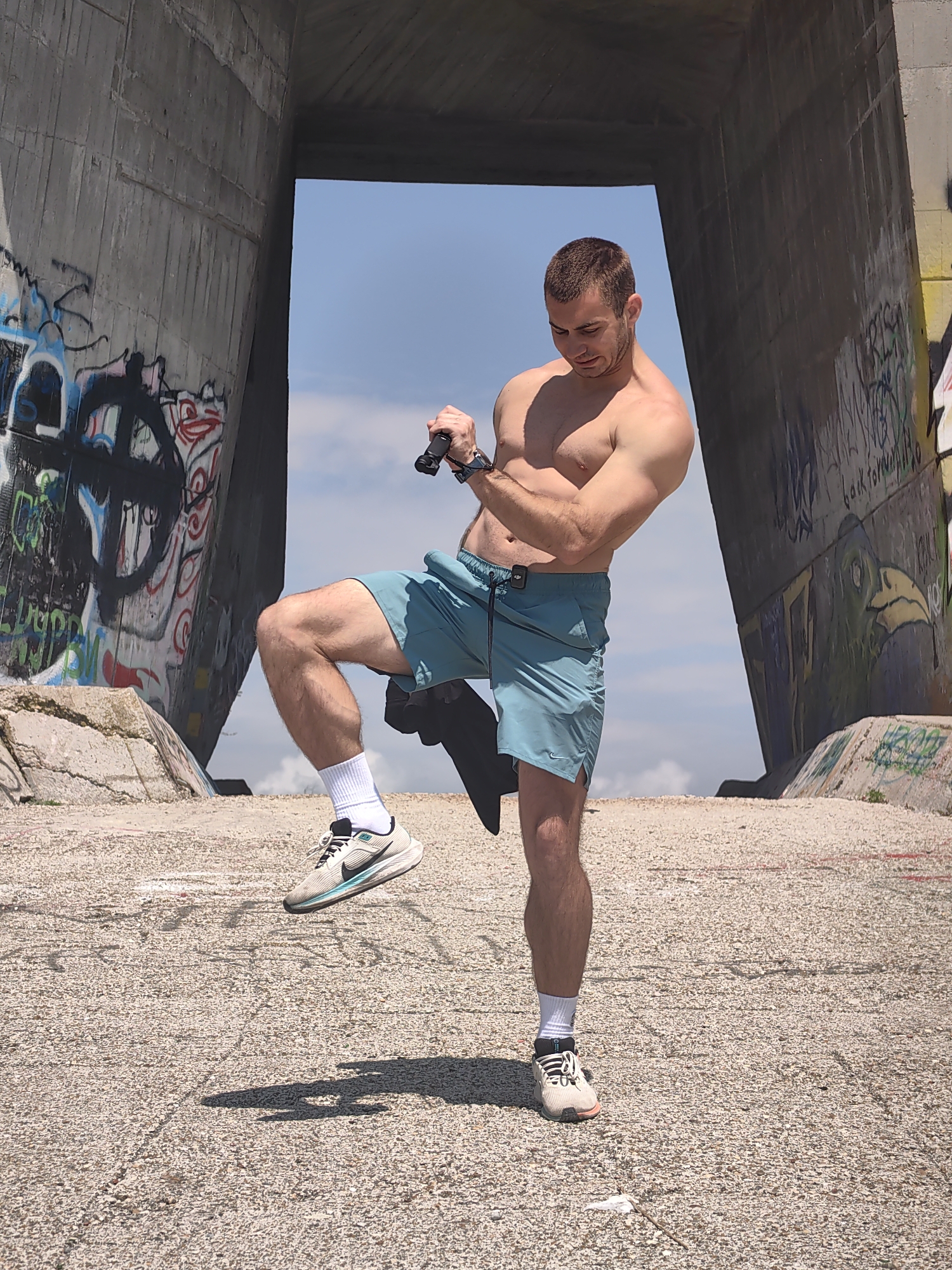 An outdoor scene in Varna, Bulgaria, depicts a shirtless male individual posing dynamically under a large concrete architectural structure. The man has short, dark hair, a muscular physique, and is wearing light teal athletic shorts with an Adidas logo, white socks, and white and black athletic shoes, with a Nike swoosh visible on his raised left foot's shoe. A dark smartwatch is visible on his left wrist. In his right hand, he holds a black, cylindrical handheld device, possibly a gimbal or camera accessory, which he appears to be looking at or adjusting, with his head tilted downwards. His left leg is bent at the knee, with the foot raised off the ground, while his right foot is firmly planted on a light-colored, pebbly ground surface. Shadows indicate bright sunlight. The prominent background feature is a massive concrete structure, likely an underpass or bridge support, forming a large rectangular opening. The interior and exterior surfaces of the concrete columns are extensively covered in colorful graffiti, featuring various tags, symbols (including a peace sign), and stylized artwork in black, red, white, blue, green, and yellow paints. The sky visible through the opening is light blue with some scattered white clouds.