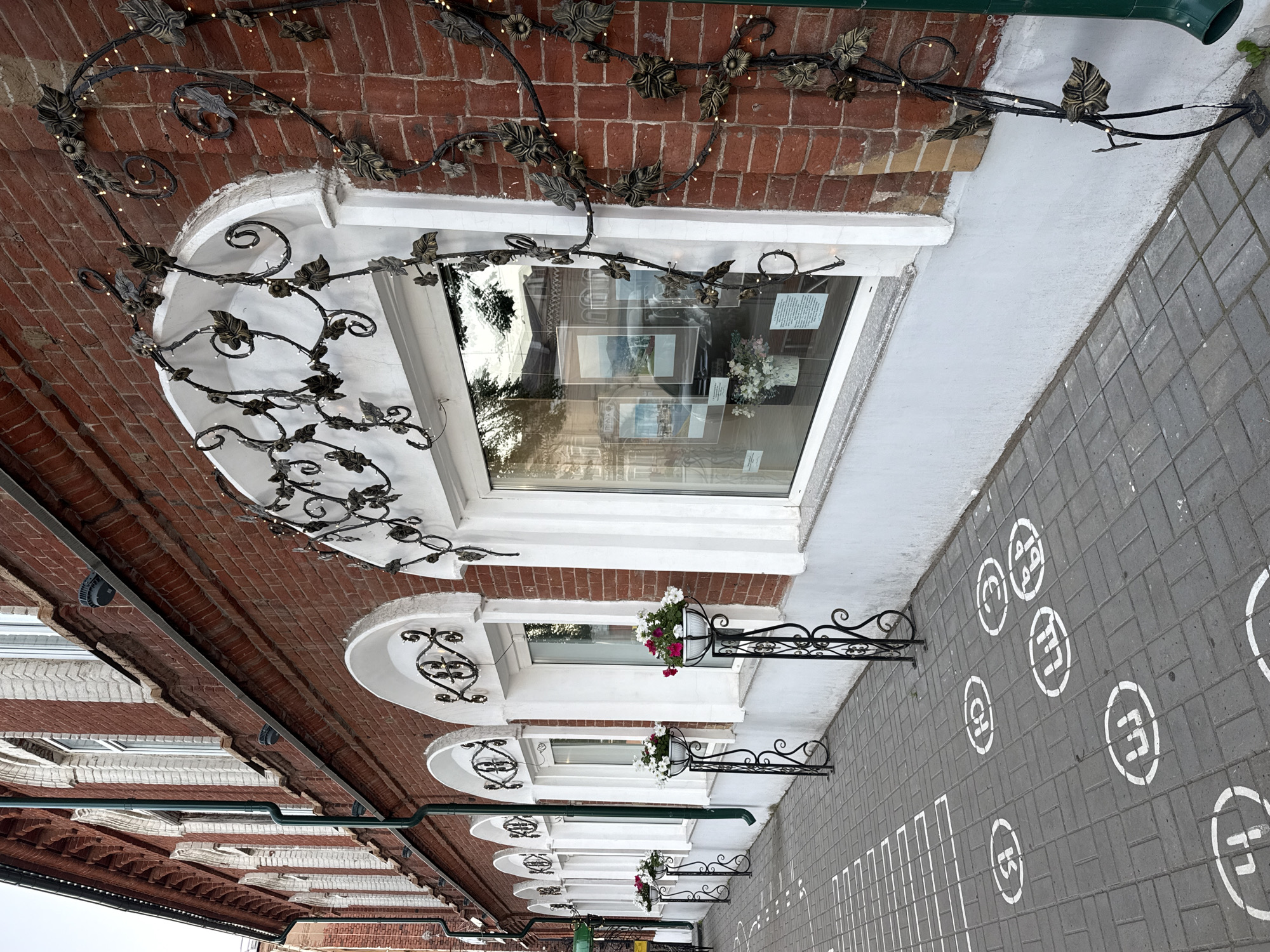 The image depicts a sidewalk with several white circular markings containing Cyrillic letters on a gray brick-paved walkway. These markings suggest a game or some form of urban art. To the right of the sidewalk is the facade of a building constructed of red brick, with a white plastered base. The building features multiple arched windows, some of which are adorned with decorative black wrought ironwork that extends upwards, twisted into vine-like shapes and decorated with metal leaves and small lights. Flower pots with pink and white flowers are placed on black metal stands in front of some of the windows. The windows themselves are white-framed and reflect the surroundings. A green drainpipe runs vertically along the side of the building. The overall scene suggests an urban street with artistic embellishments and pedestrian-oriented markings. The location is identified as Votkinsk, Russia. There are no people visible in the image.