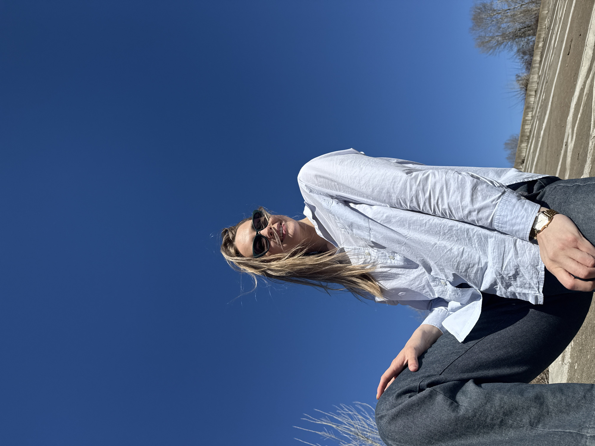 A woman is positioned on what appears to be a concrete structure. She wears sunglasses, a light-colored button-down shirt, and dark pants. Her blonde hair is loose, falling towards the ground. She is also wearing a gold watch. The background features a clear blue sky, and hints of bare trees and the edge of a concrete structure are visible. This photo was taken in Chaikovsky, Russia.