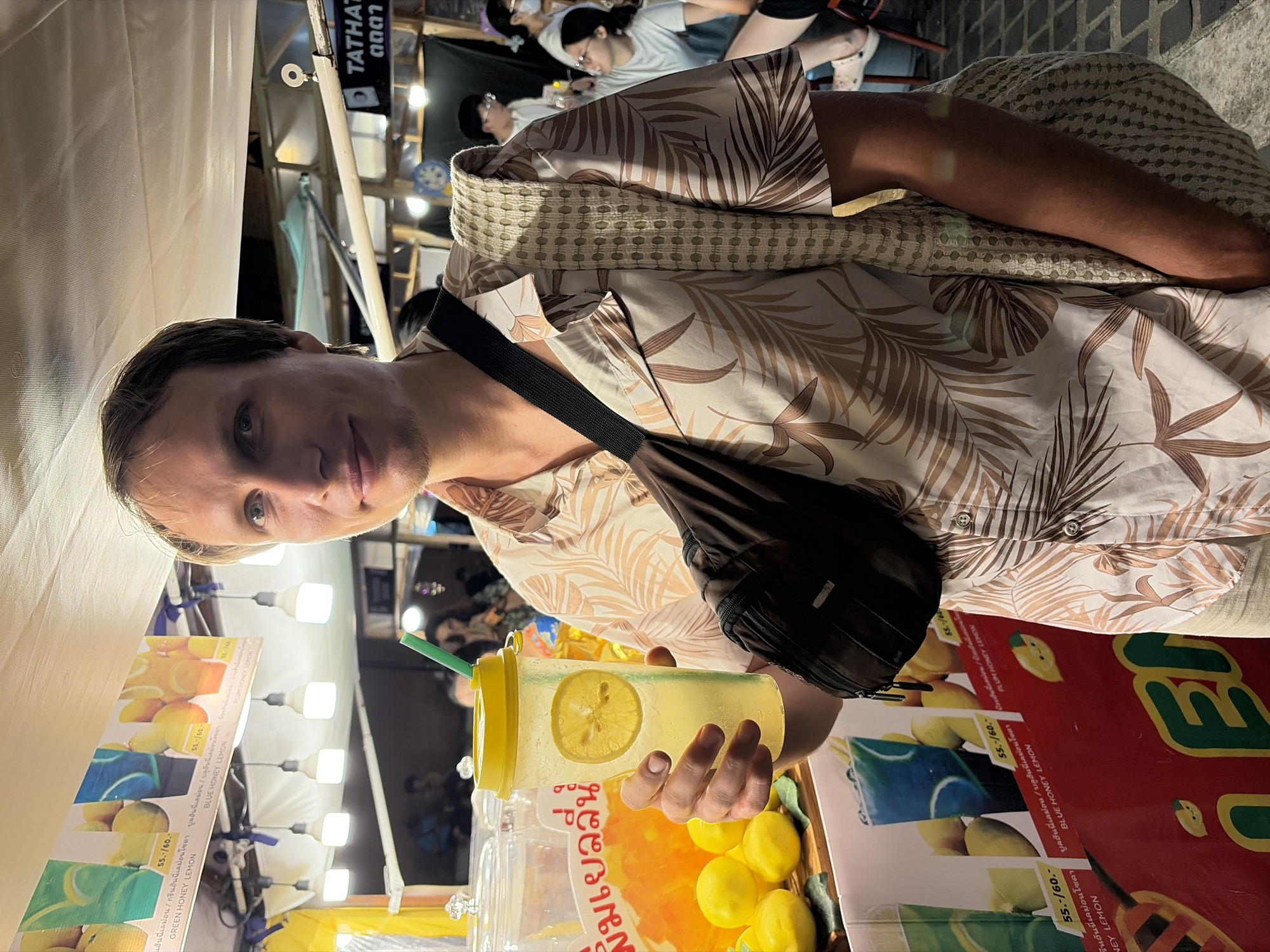 A man stands at a fruit juice stand in Chiang Mai City Municipality, Thailand, holding a cup of lemonade with a slice of lemon. He wears a tropical-print shirt and a crossbody bag. Behind him, various fruit juices and lemons are displayed, and other customers can be seen in the background. The scene appears to be an outdoor market or street food stall at night, with artificial lighting illuminating the area.